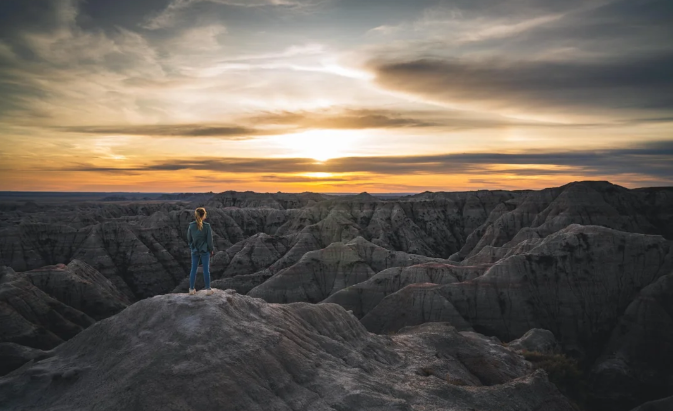A person stands on a rocky hilltop overlooking rugged, eroded badlands at sunset, with dramatic clouds and warm light filling the sky—a breathtaking scene reminiscent of vistas found in American National Parks.