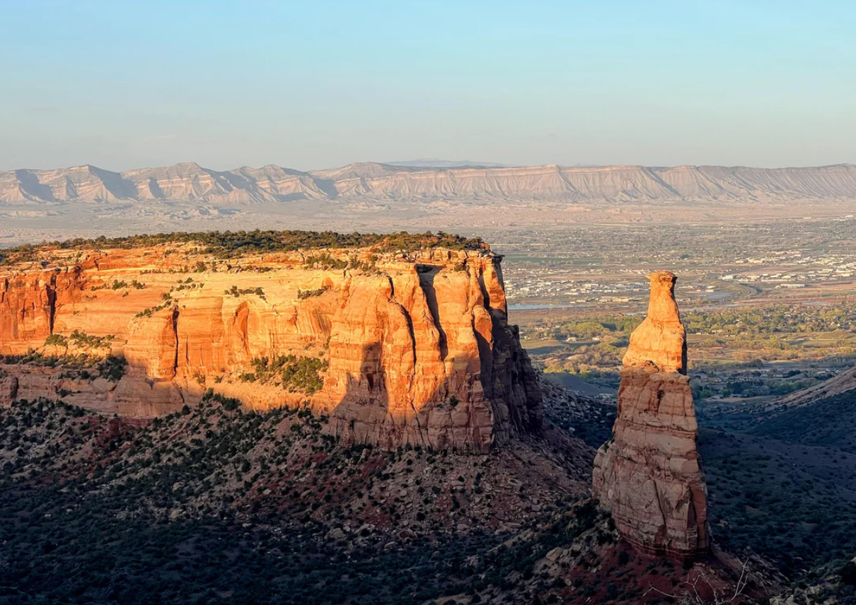 Red sandstone cliffs and a tall spire rise from a desert landscape within one of the iconic American National Parks, bathed in golden sunlight, with distant mesas and a broad valley visible under a clear blue sky.