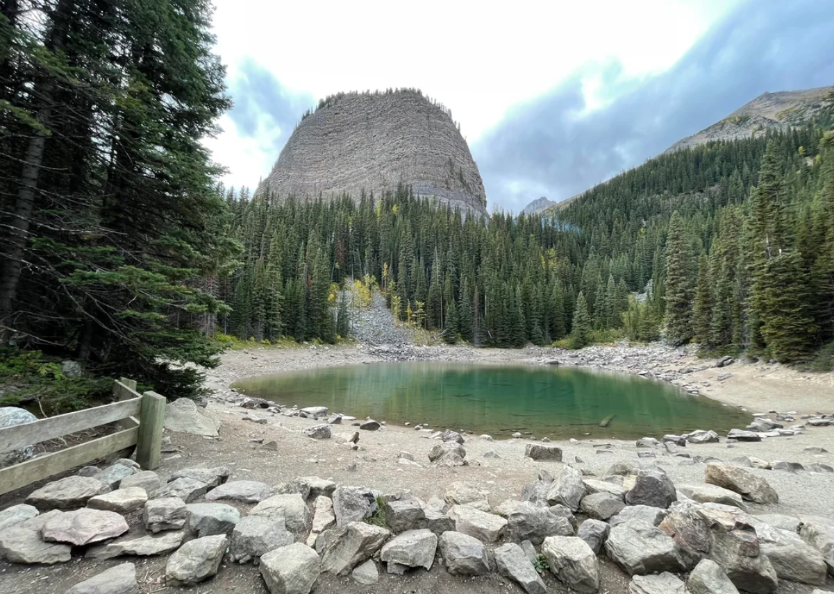 A small, circular lake surrounded by rocks and pine trees sits below a large, rounded mountain under a cloudy sky—scenery reminiscent of the breathtaking landscapes found in American National Parks. A wooden fence is visible in the left foreground.