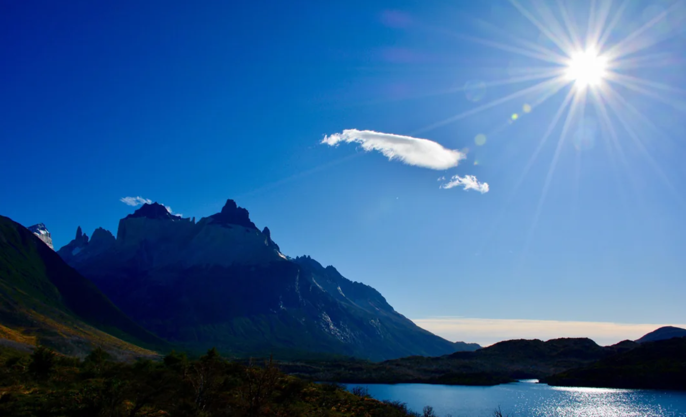 Mountain peaks rise beneath a bright blue sky, with a single white cloud floating and the sun shining above—an iconic scene reminiscent of American National Parks. A lake at the base of the mountains reflects the sunlight, capturing the breathtaking beauty found throughout these treasured landscapes.