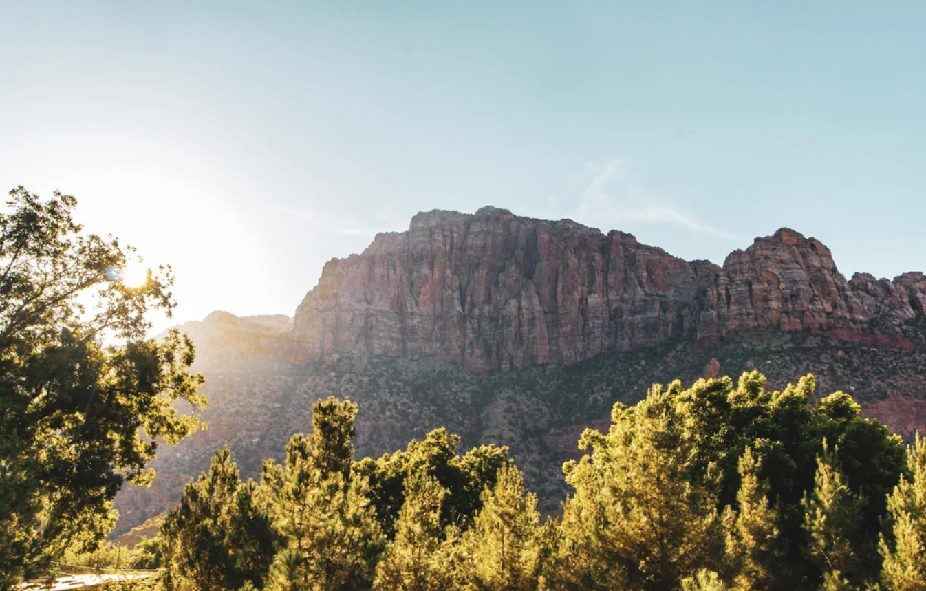 The sun rises behind tall red rock cliffs and green trees under a clear blue sky, creating a bright and scenic landscape reminiscent of the breathtaking vistas found in American National Parks.