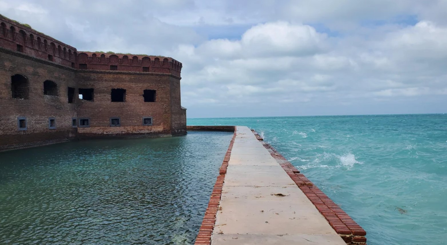 A brick walkway leads to a historic brick fort, one of the hidden gems found within American National Parks, surrounded by calm blue-green water under a partly cloudy sky. Waves crash against the right side of the fort.