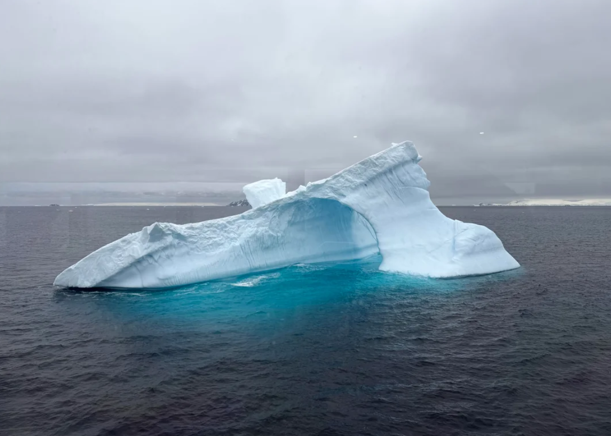 A large, jagged iceberg floats in dark ocean water under a cloudy sky, with a vivid blue area visible below the surface and a distant icy coastline on the horizon.