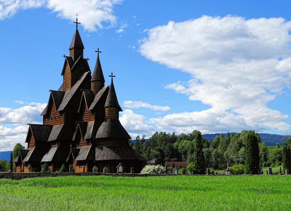 Wooden stave church with multiple steep roofs and crosses, set in a green field with trees, under a blue sky with scattered clouds.