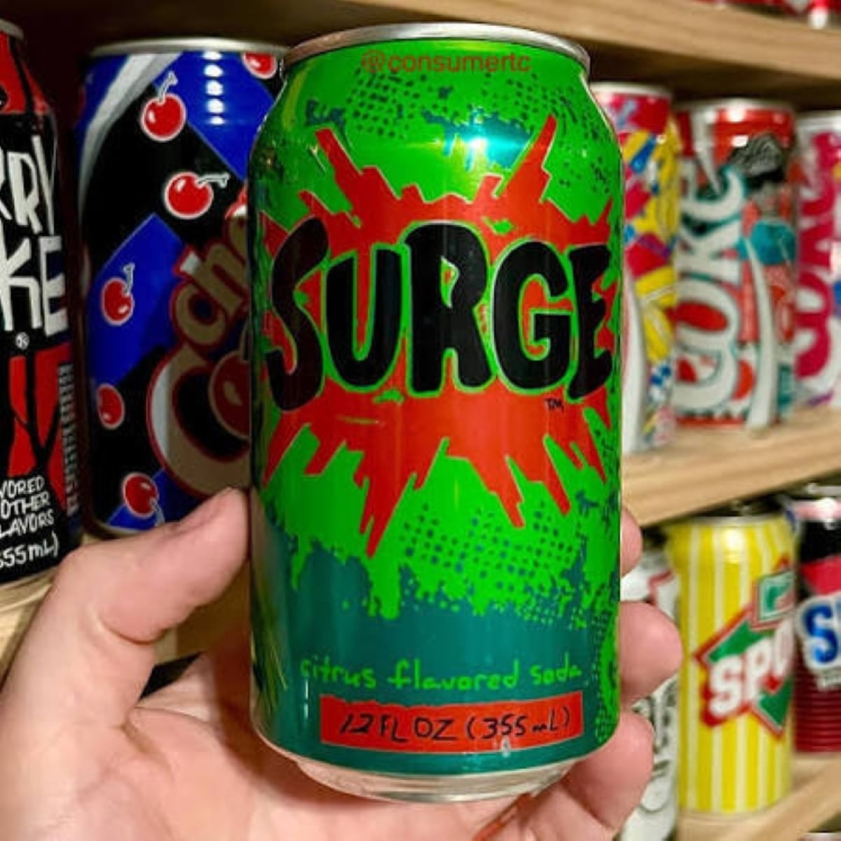 A hand holds a can of Surge citrus-flavored soda in front of shelves lined with various colorful Soda Pops cans. The Surge can is green with a red burst design and bold black lettering.