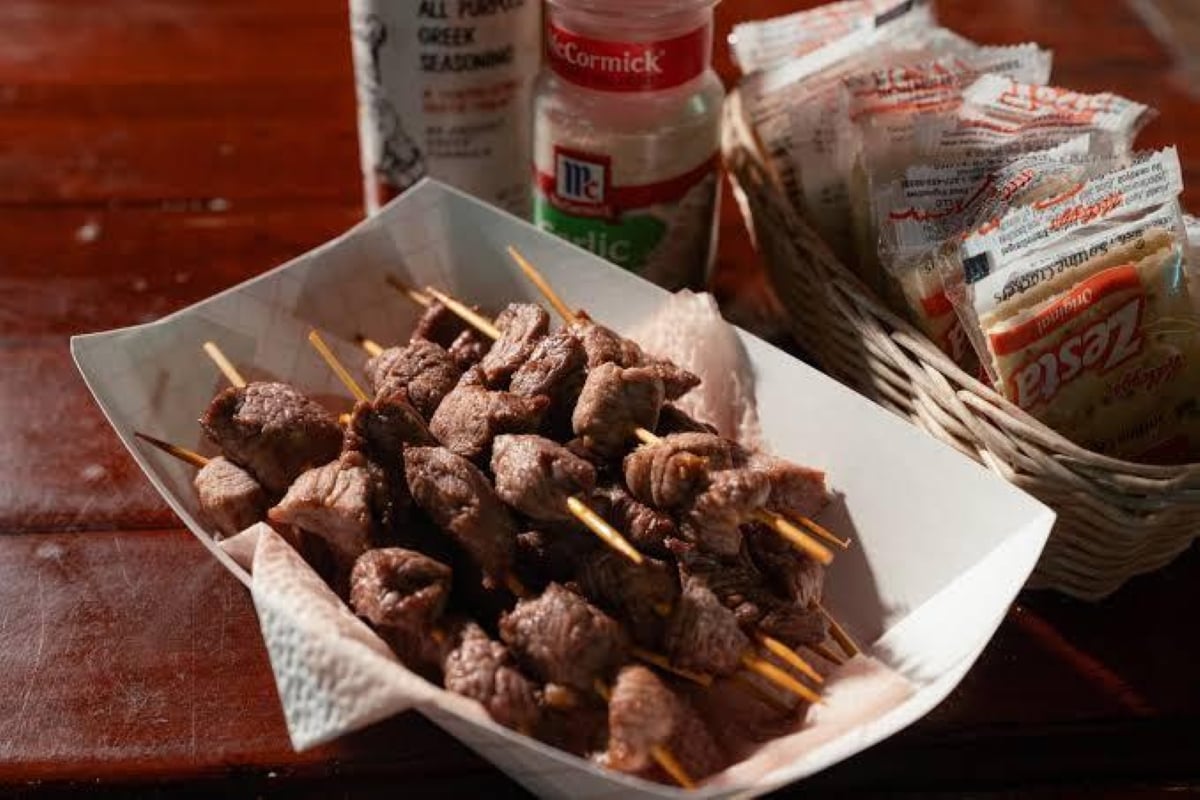 A paper tray holds skewers of grilled meat on a wooden table, with seasoning bottles and a basket of assorted condiment packets in the background.
