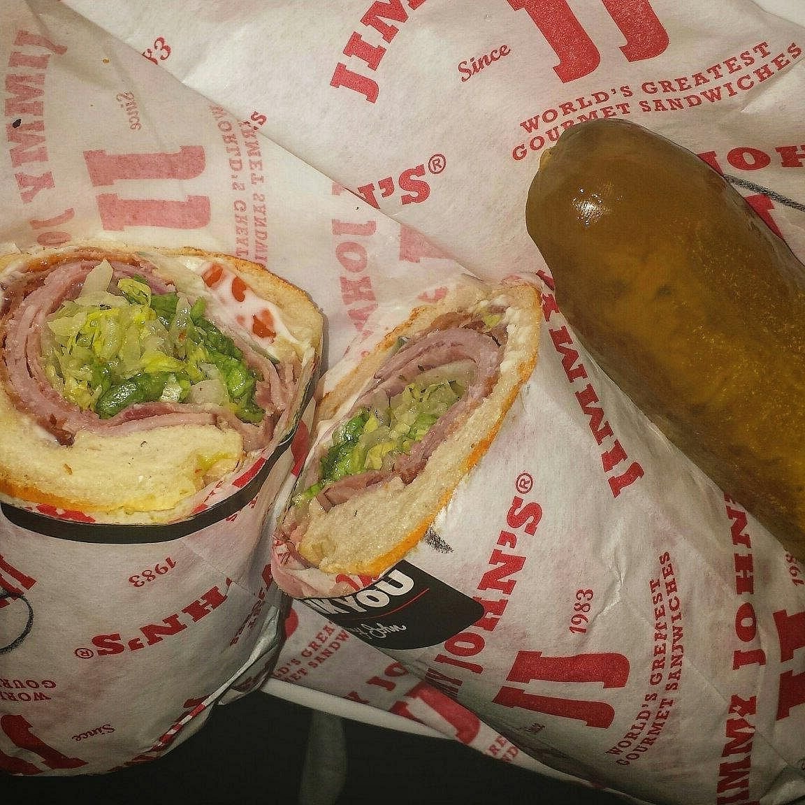 A sliced Jimmy John’s sub sandwich filled with meats, cheese, lettuce, and sauce is shown alongside wrapped sandwiches and a whole pickle.