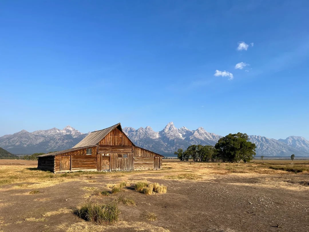 A wooden barn stands on dry grass with a backdrop of distant mountains under a clear blue sky, evoking the quiet charm often discovered on scenic road trips.