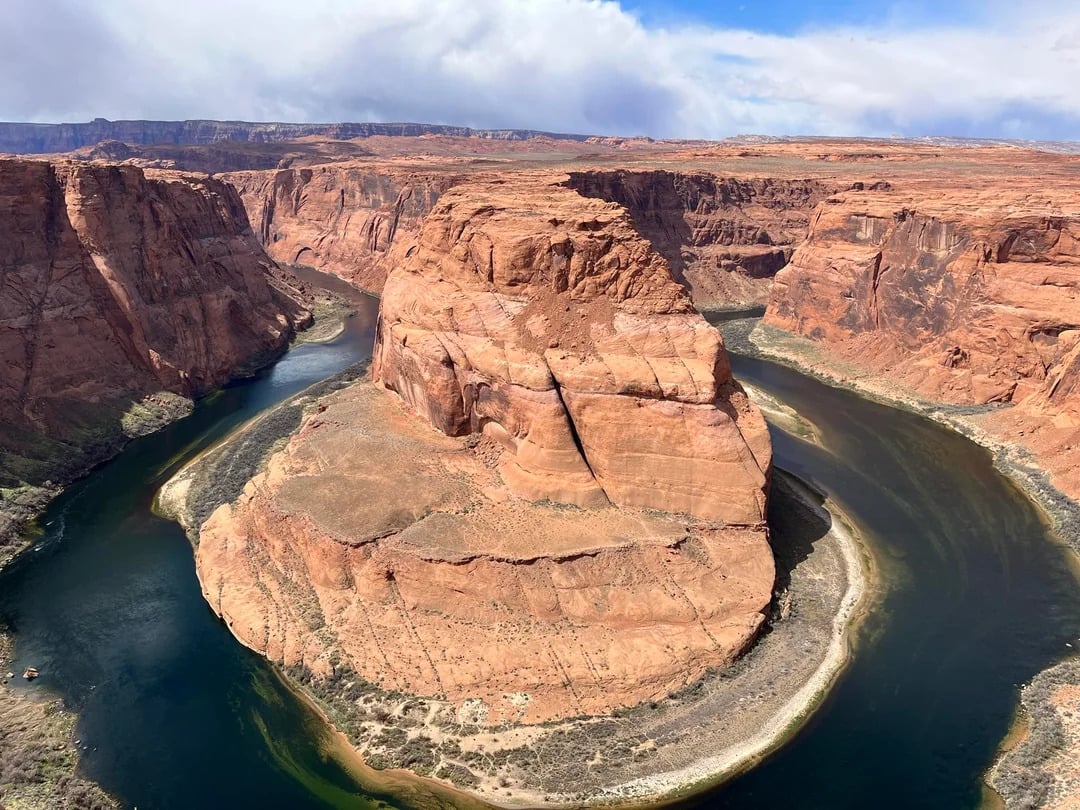 Aerial view of Horseshoe Bend, a dramatic meander in the Colorado River surrounded by steep, orange-brown canyon walls under a partly cloudy sky—a must-see highlight for memorable road trips through the American Southwest.