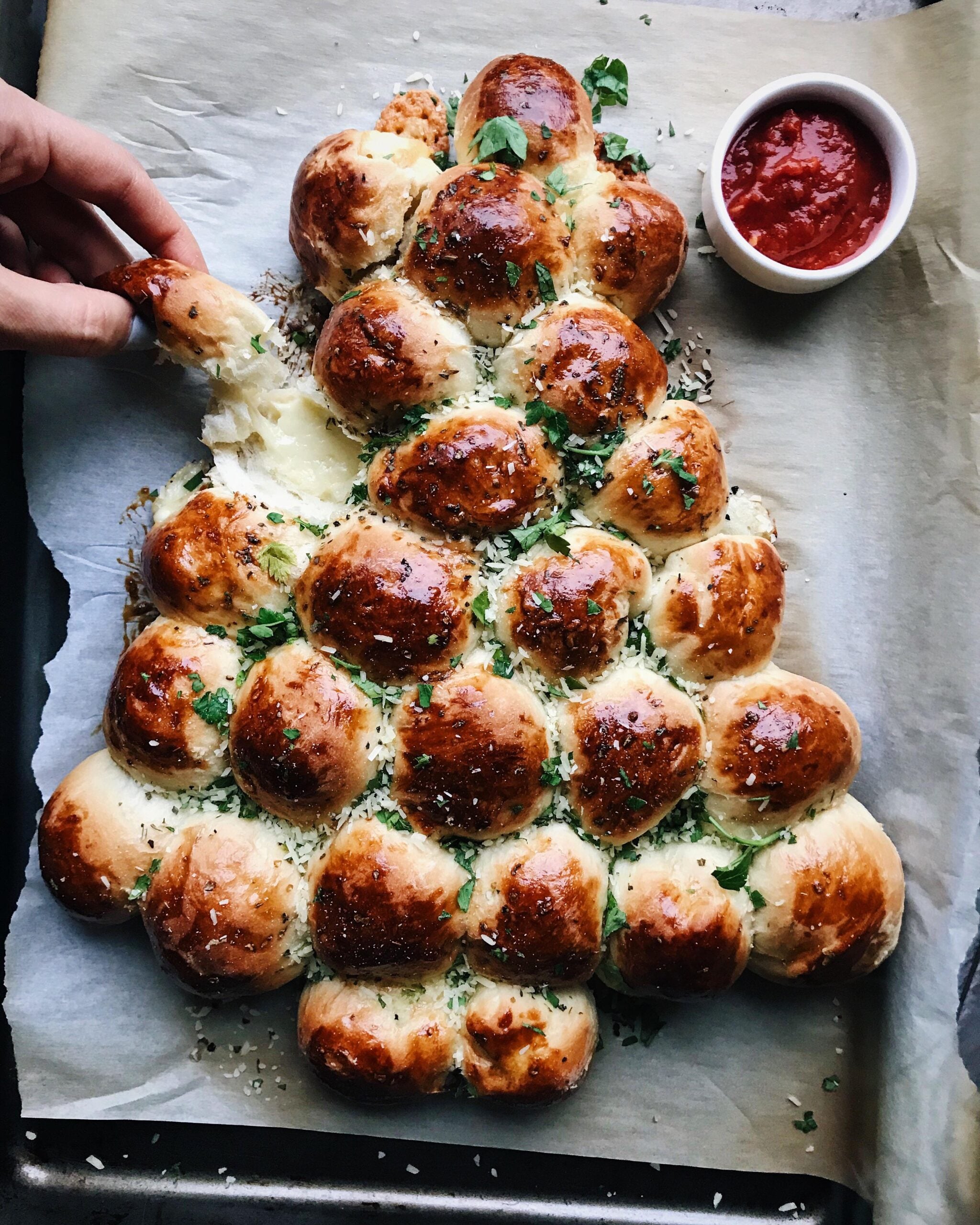 Tree-shaped pull-apart bread made of golden rolls topped with herbs, with a small bowl of red dipping sauce nearby. A hand pulls off a piece from the left side.