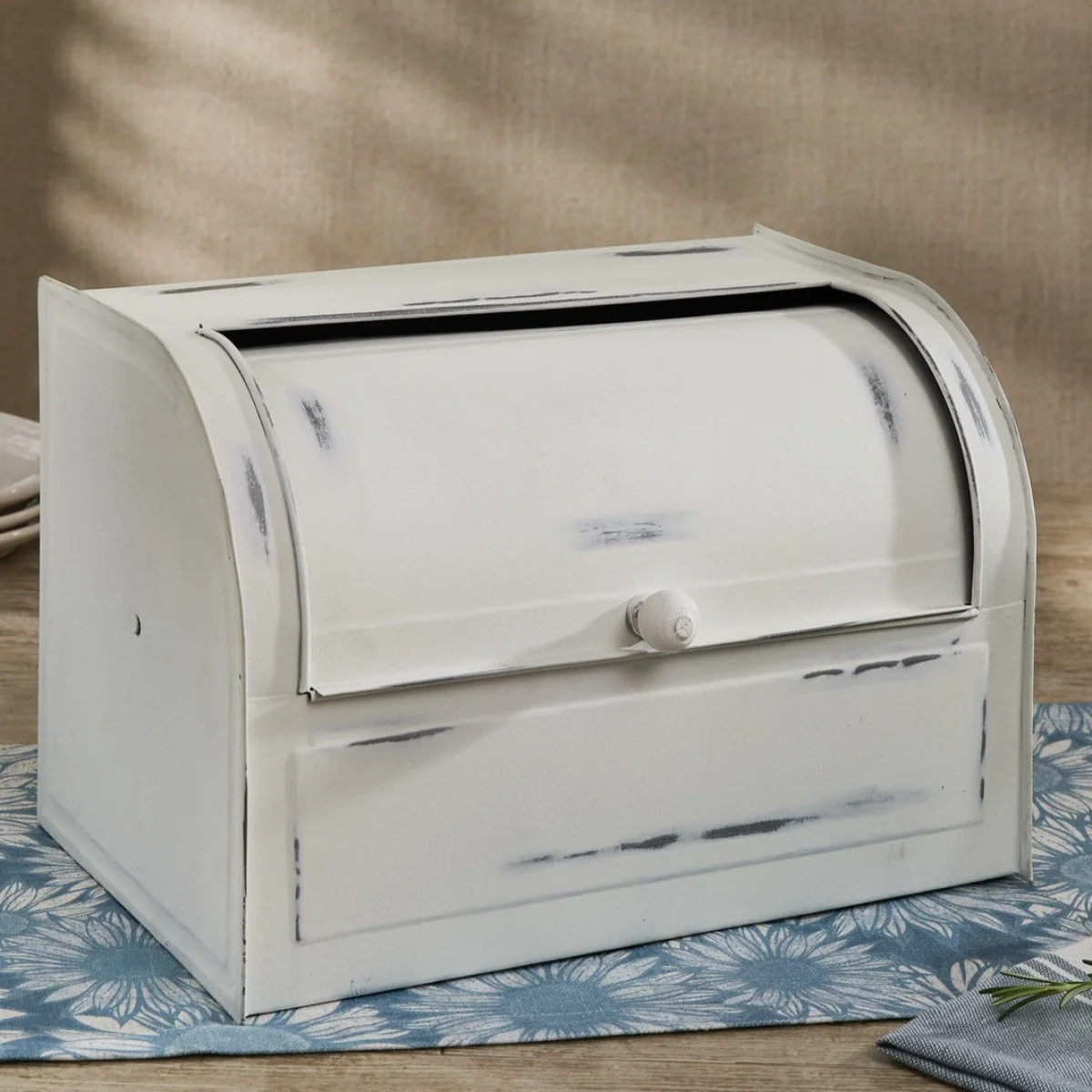 A white vintage-style bread box with a roll-top lid sits on a blue floral placemat on a wooden surface against a neutral background.