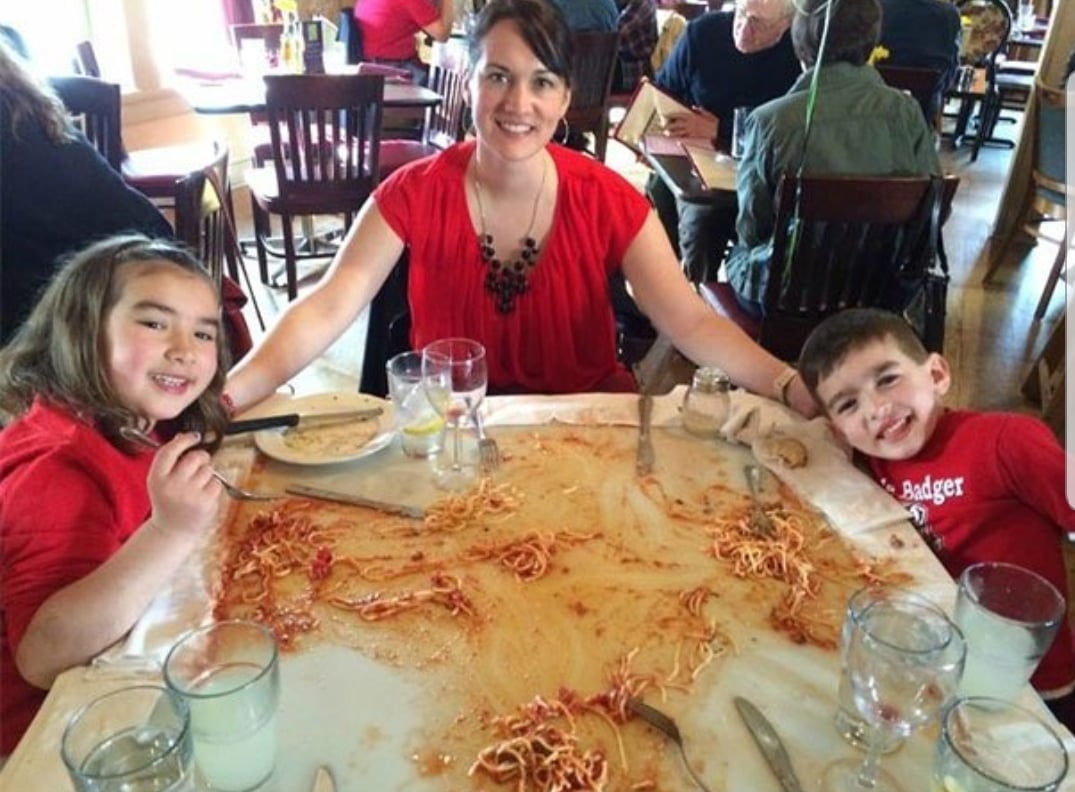 A woman and two kids smile at a restaurant table covered in messy, spilled spaghetti. Plates are empty, and glasses of water are on the table. All three wear red tops and appear to be enjoying themselves.