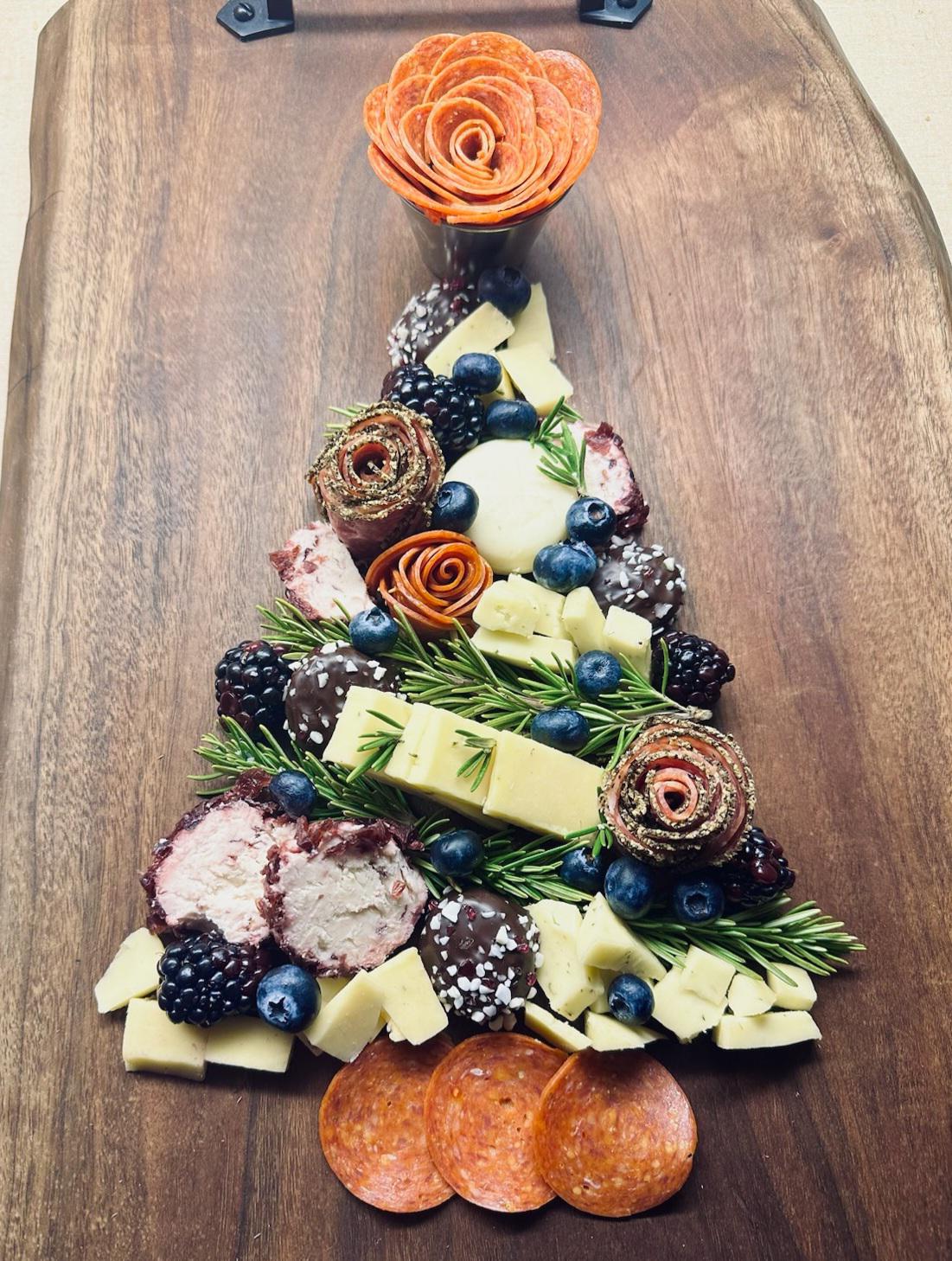 A charcuterie board arranged in the shape of a Christmas tree, featuring cheese cubes, meats, berries, rosemary sprigs, and a salami rose at the top—all on a wooden board—makes for festive homemade Christmas treats sure to delight your guests.