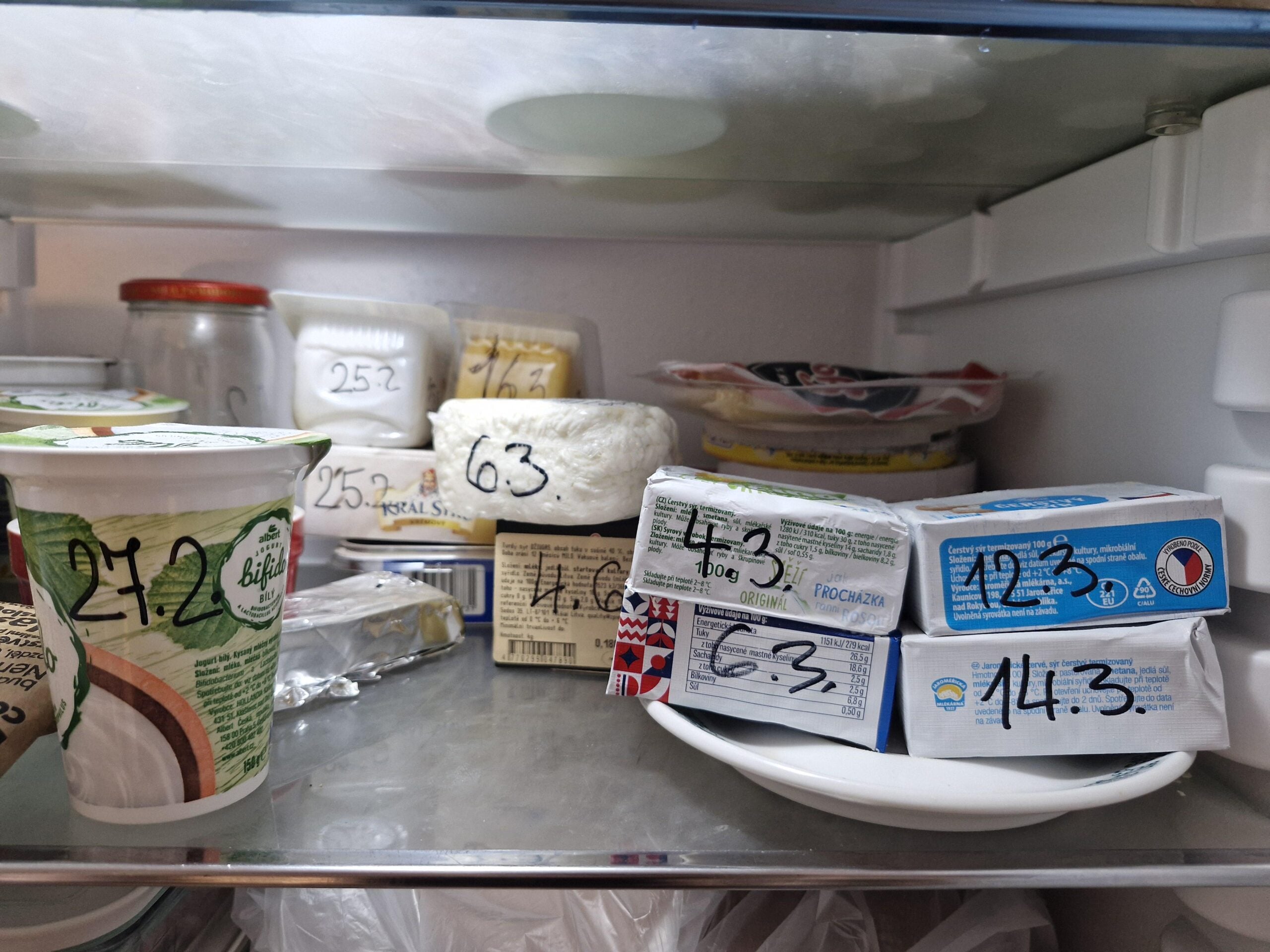 Various dairy products, including yogurt and cheese, are stored on a refrigerator shelf. Several items are labeled with handwritten dates on them, possibly indicating expiration or opened dates.