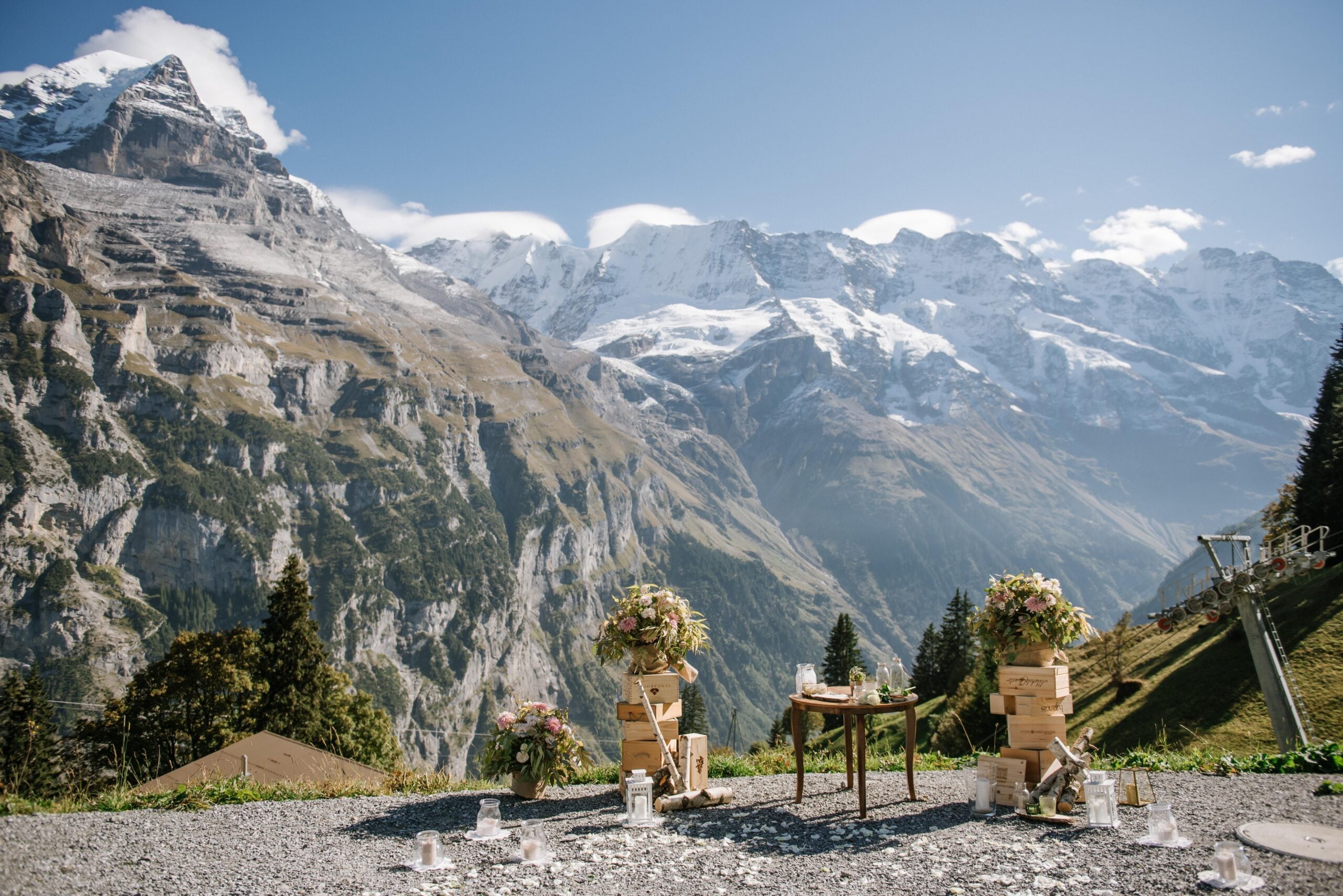 Outdoor wedding setup on a mountain, with two flower-decorated chairs and a small table. Snow-capped peaks and a blue sky create a scenic alpine backdrop.