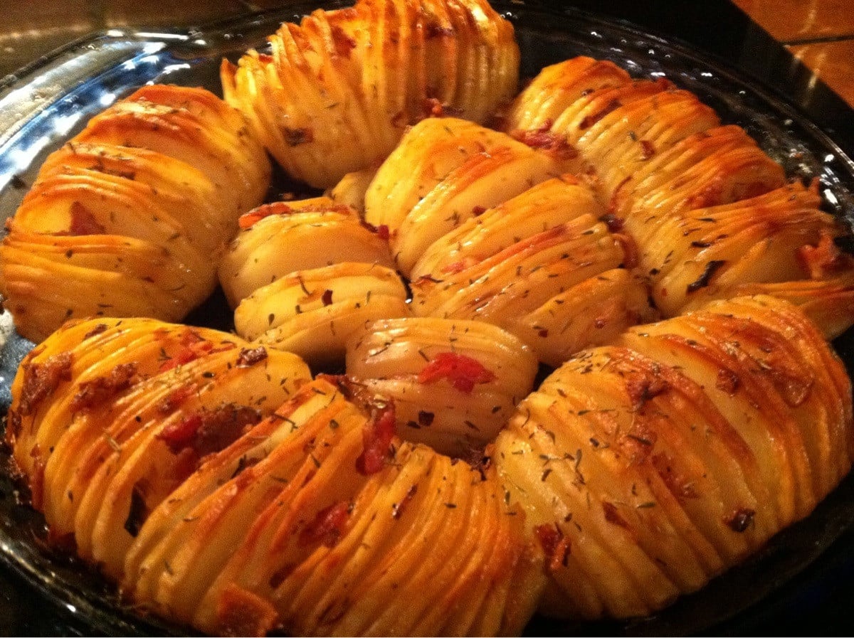 A close-up of Hasselback potatoes, thinly sliced and baked until golden, topped with herbs and bits of bacon, served on a dark plate.