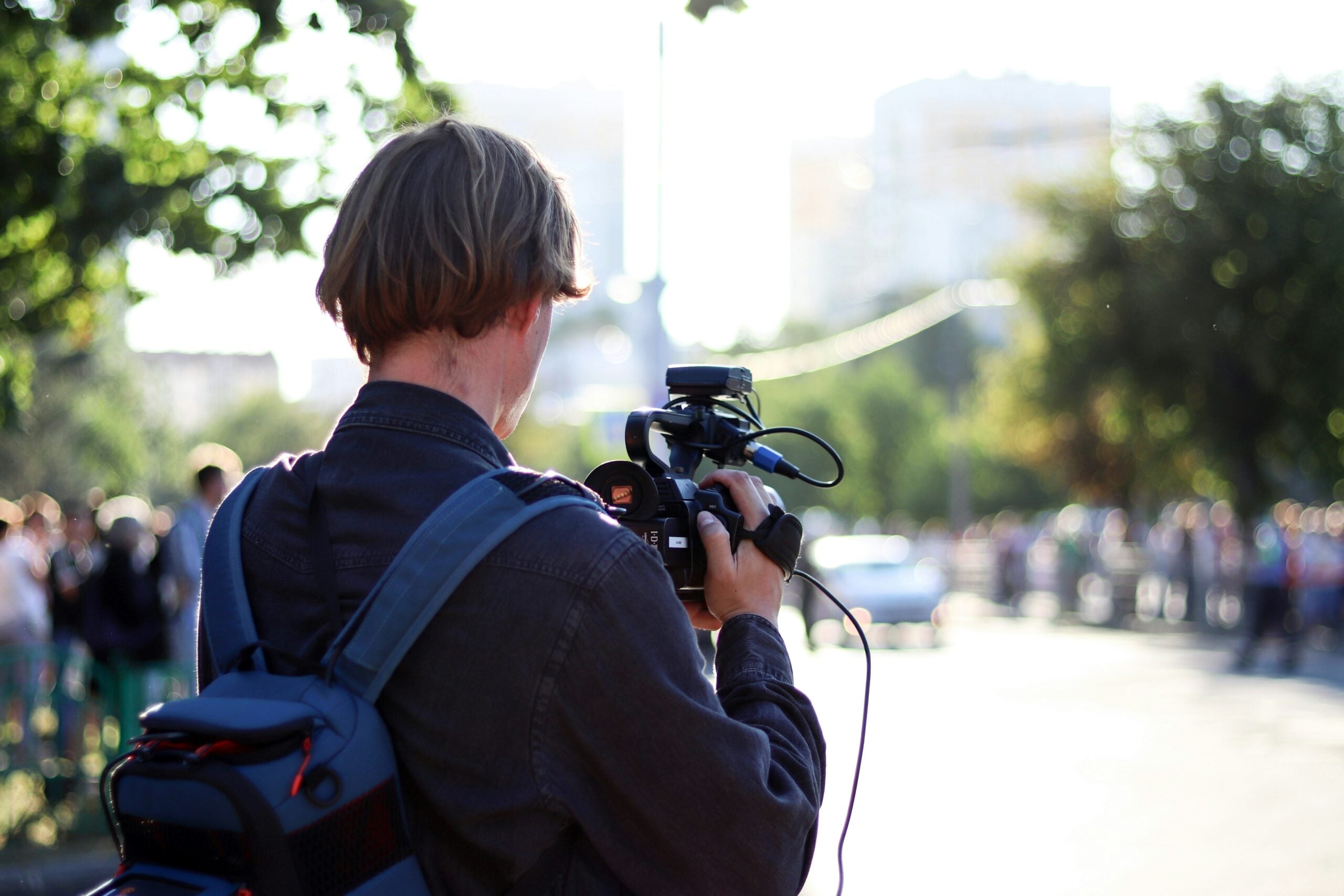 A person with short hair and a backpack films with a video camera outdoors, facing a blurred crowd and bright sunlight in the background, capturing moments that reflect life in 2026 problems.