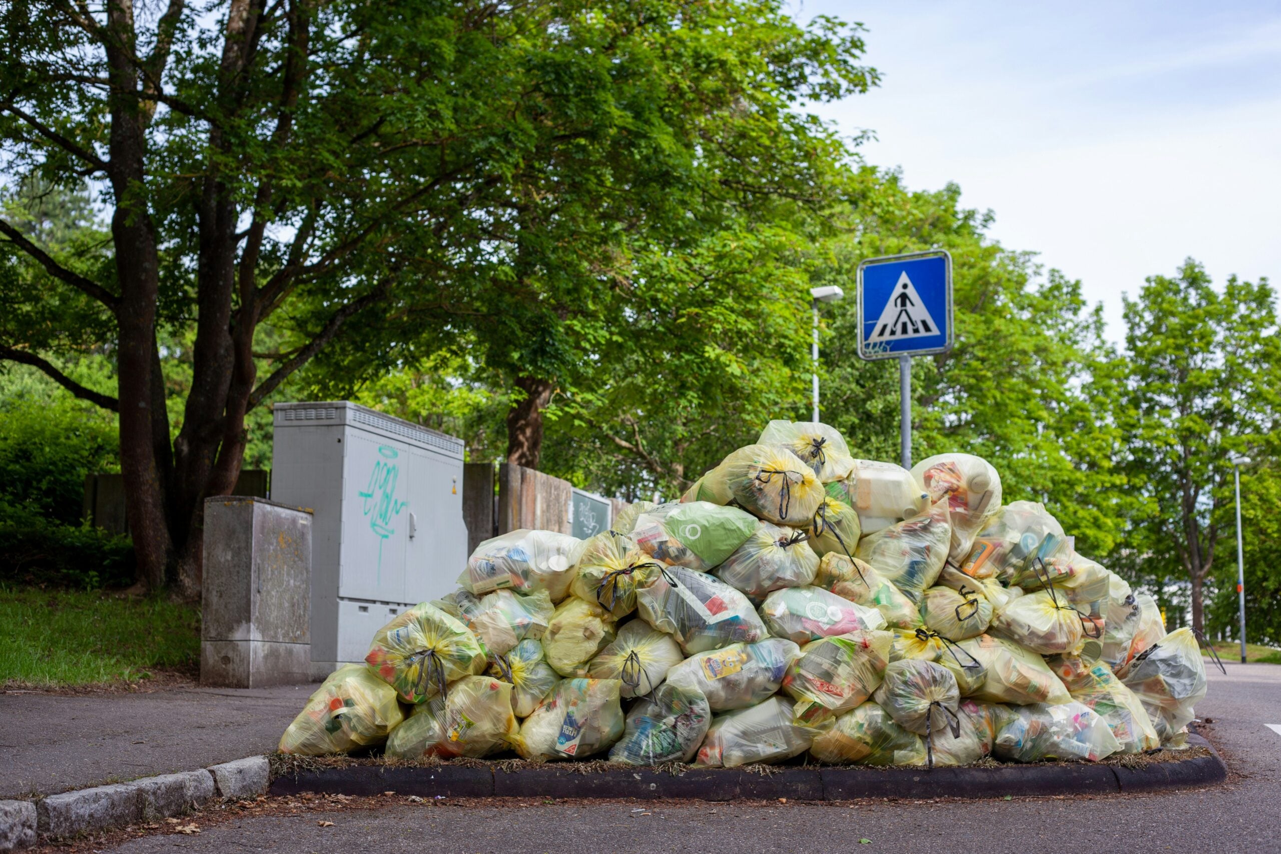 A large pile of yellow garbage bags is stacked on the side of a road near a pedestrian crossing sign and trees, highlighting some of the life in 2026 problems faced by urban communities.