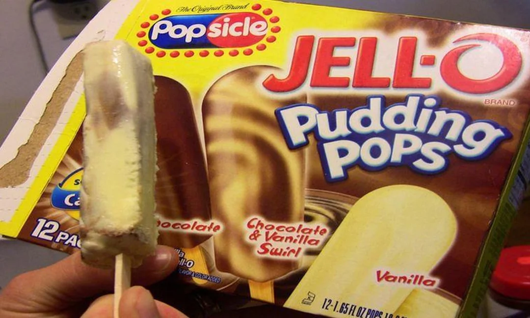 A hand holds a Jell-O Pudding Pop in front of a box showing chocolate, vanilla, and chocolate-vanilla swirl flavors. The box has Popsicle and Jell-O branding.