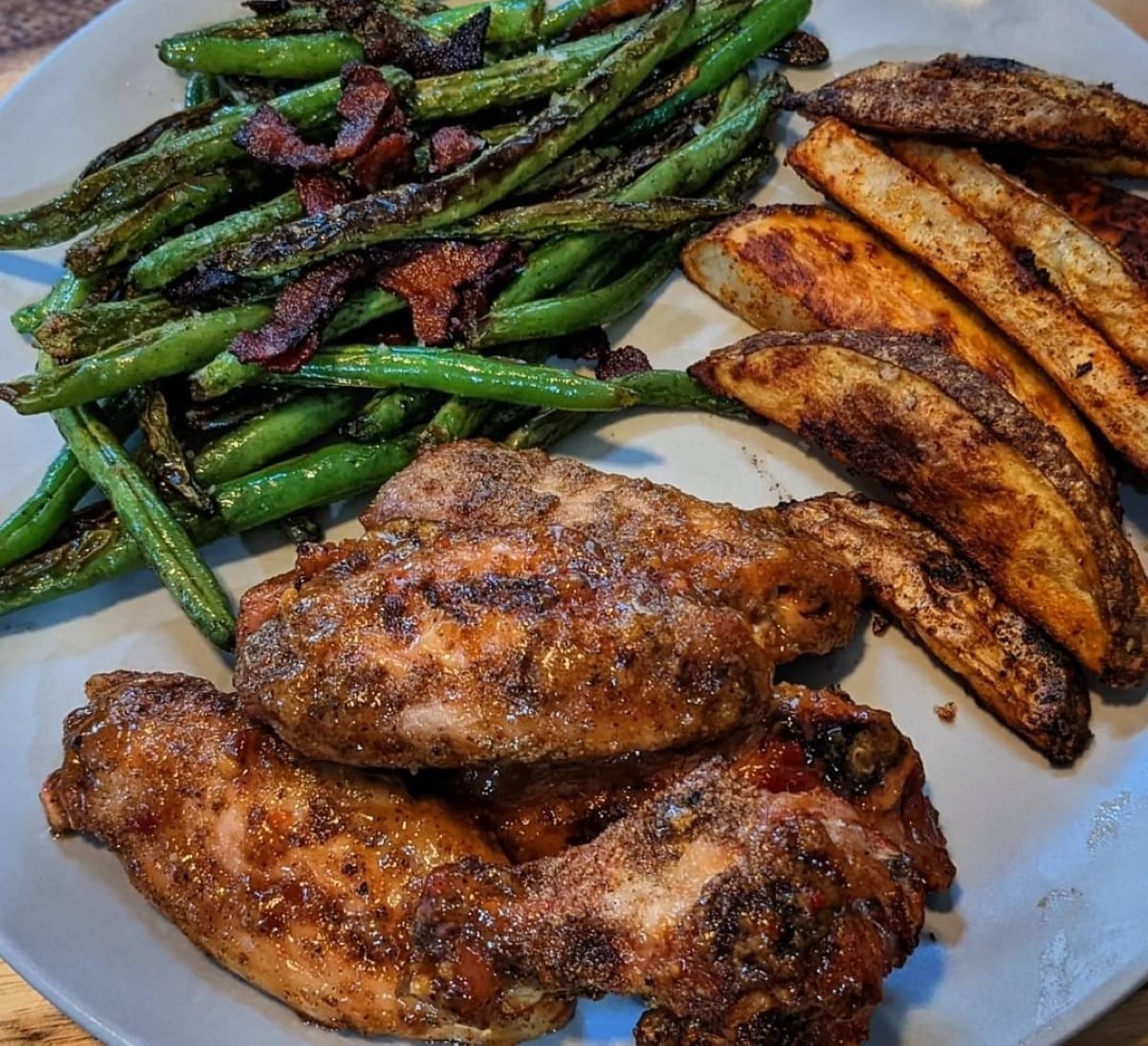 A plate with roasted chicken wings, crispy potato wedges, and sautéed green beans topped with bacon bits.