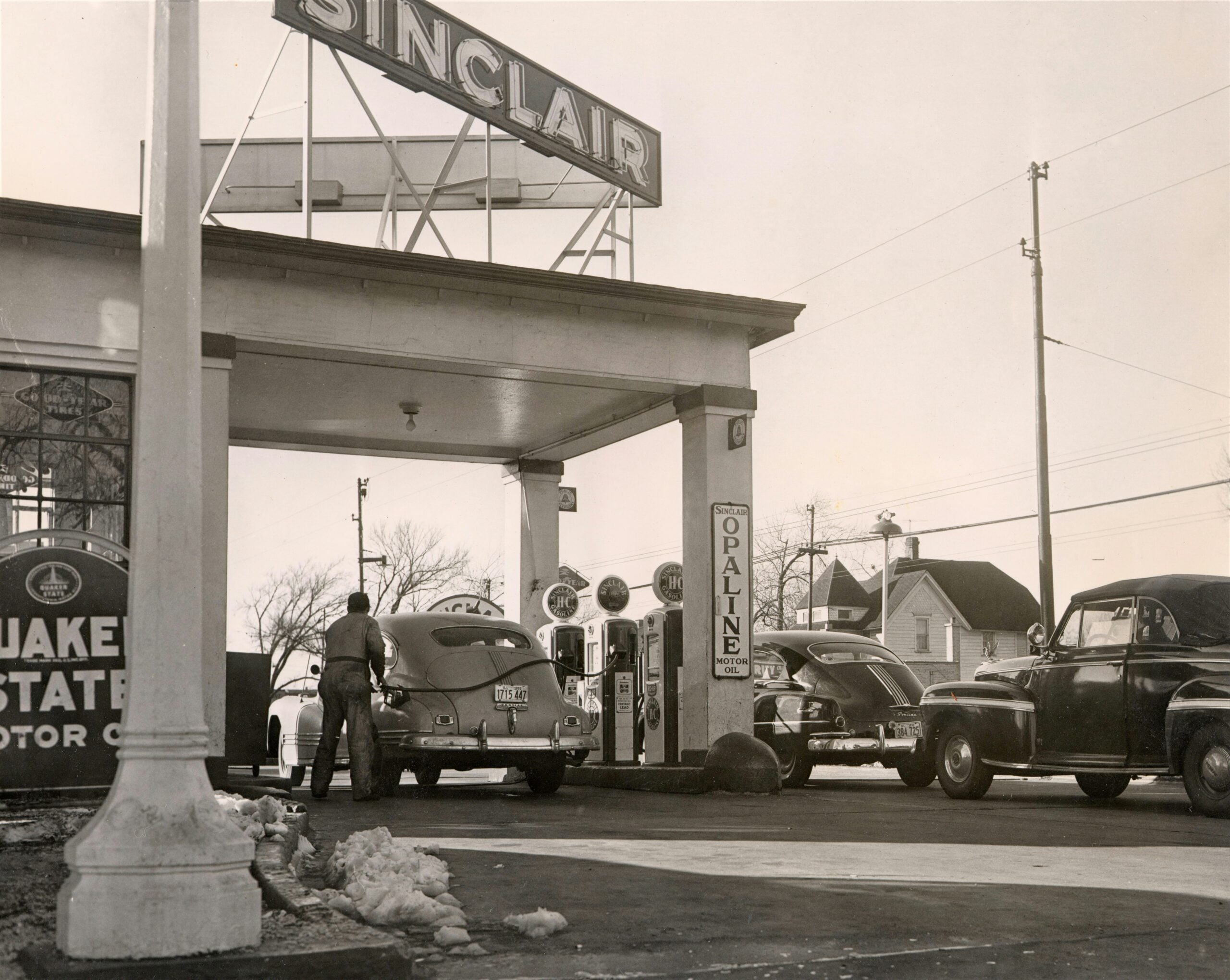 Black-and-white photo of a vintage Sinclair gas station, with three classic cars parked near gas pumps. A man is fueling a car. There are old signs, a few houses, and trees in the background.