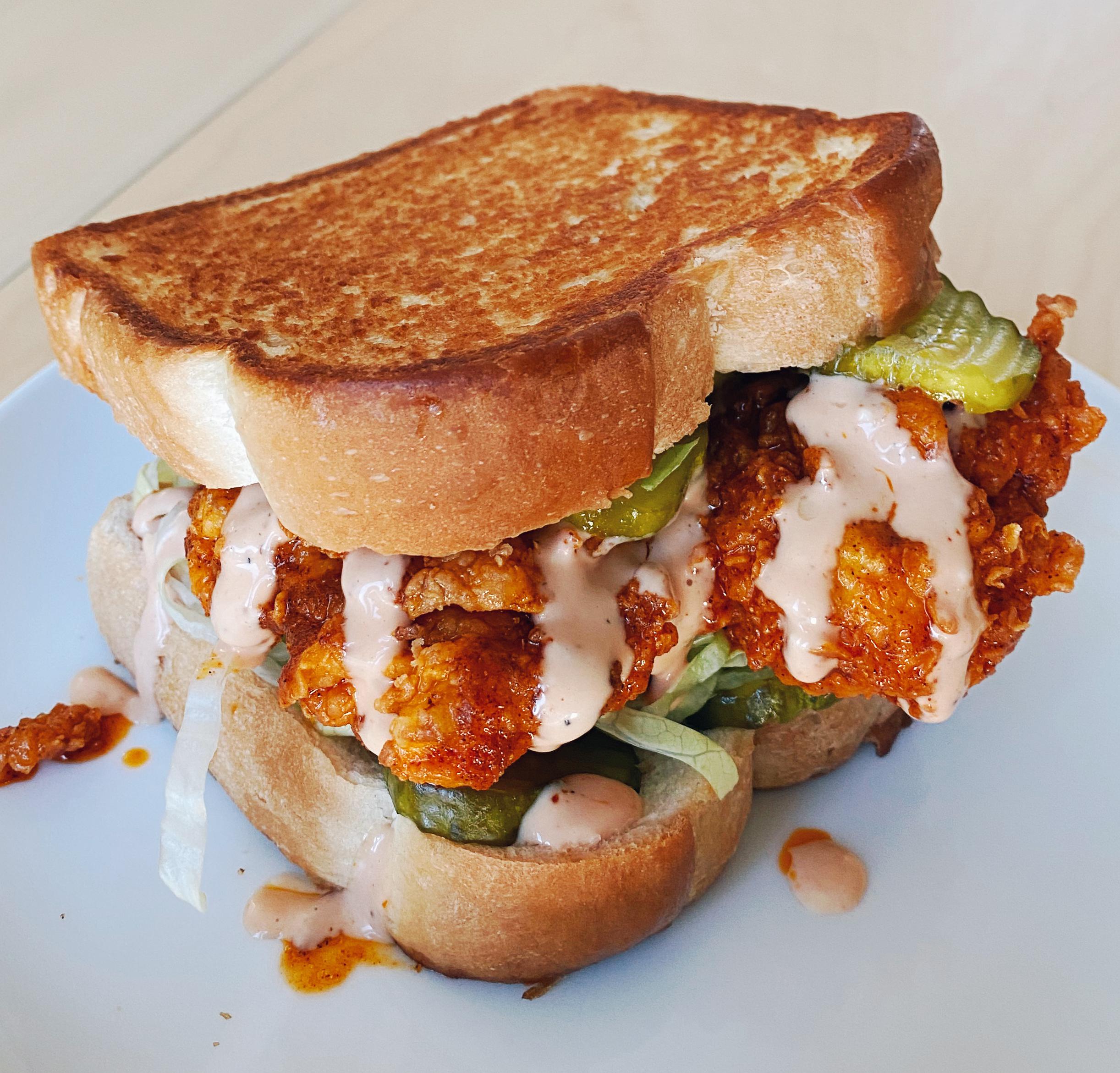A crispy fried chicken sandwich with pickles, lettuce, and creamy sauce on toasted bread, served on a white plate.