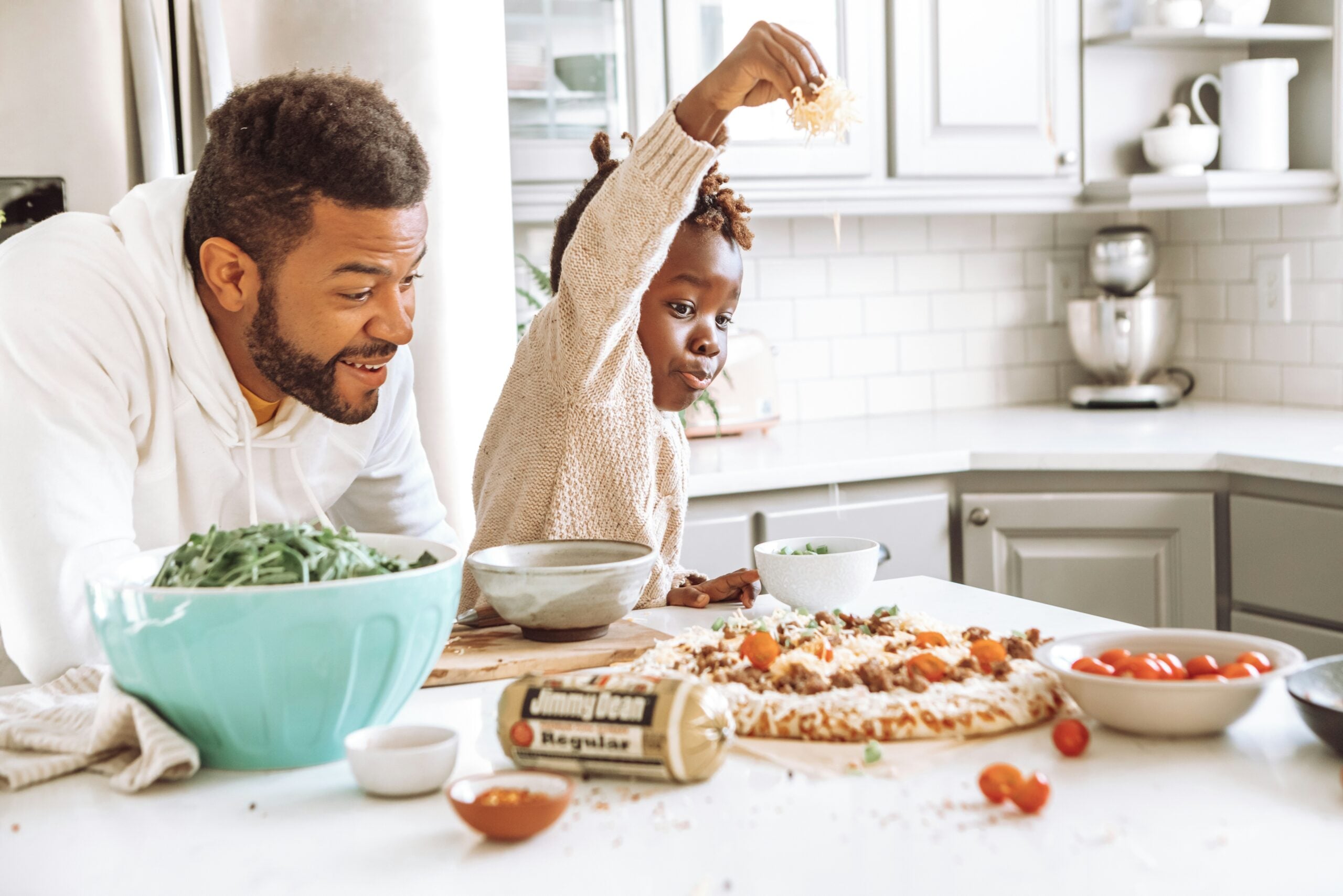 A man and child are in a bright kitchen, making pizza together. The child sprinkles cheese on a pizza, while the man smiles. Vegetables and ingredients are on the counter.