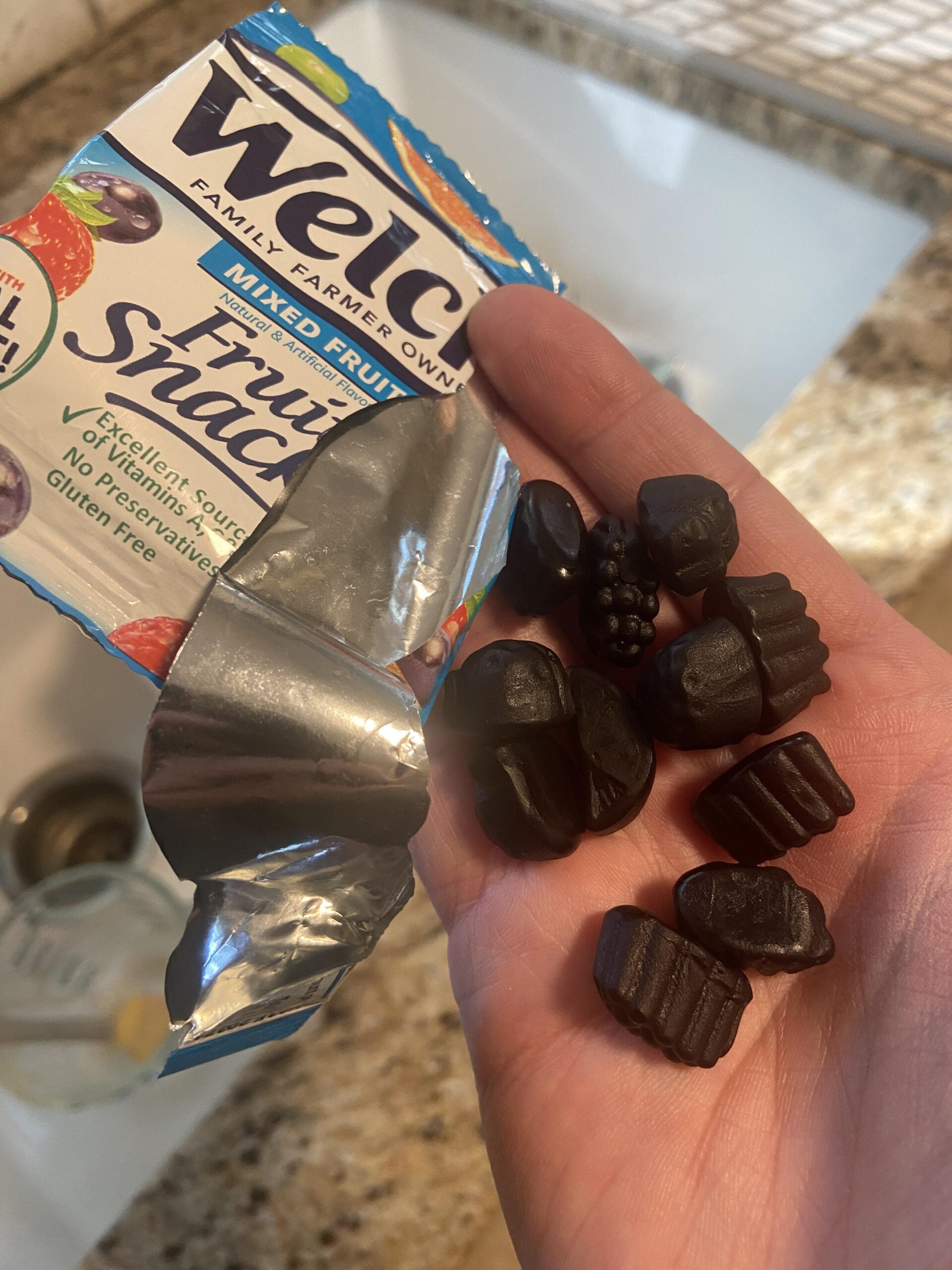 A hand holds an open Welch’s Mixed Fruit Fruit Snacks pouch and several dark gummy fruit snacks in assorted shapes above a kitchen sink.