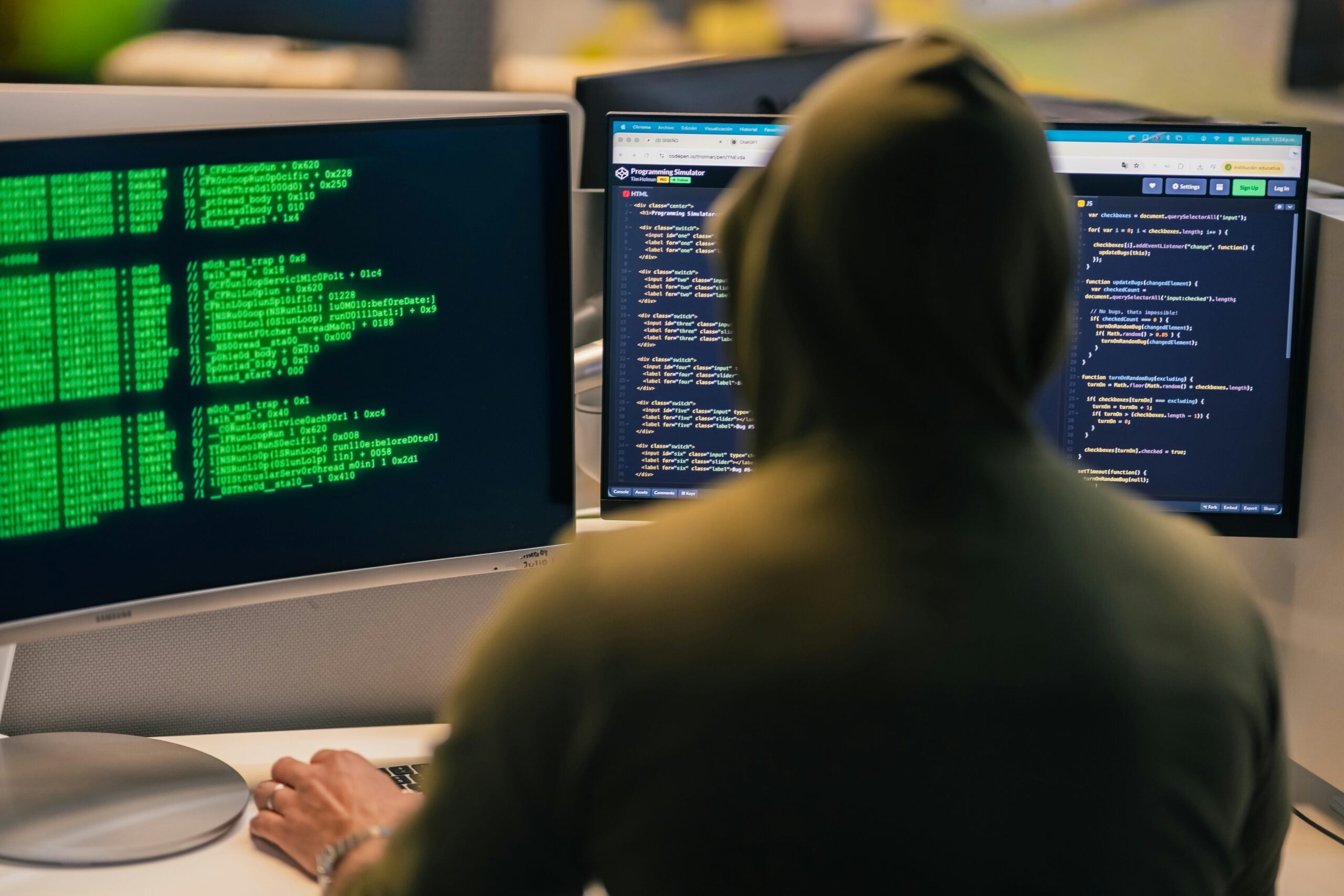 A person in a hoodie sits at a desk with two monitors displaying code, one with green text and the other with programming scripts, offering a glimpse into life in 2026 where cybersecurity and hacking activity are increasingly central to daily routines.