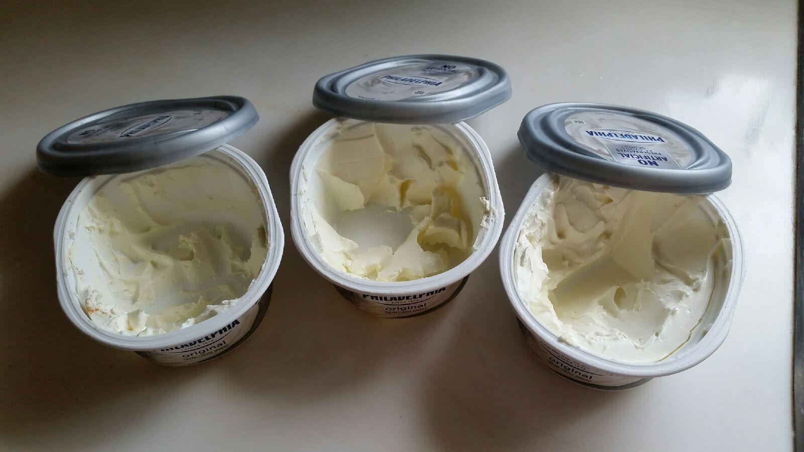 Three open tubs of Philadelphia cream cheese are shown on a surface, each partially empty with uneven scoops taken from the inside. The silver lids are peeled back and resting on top of each container.