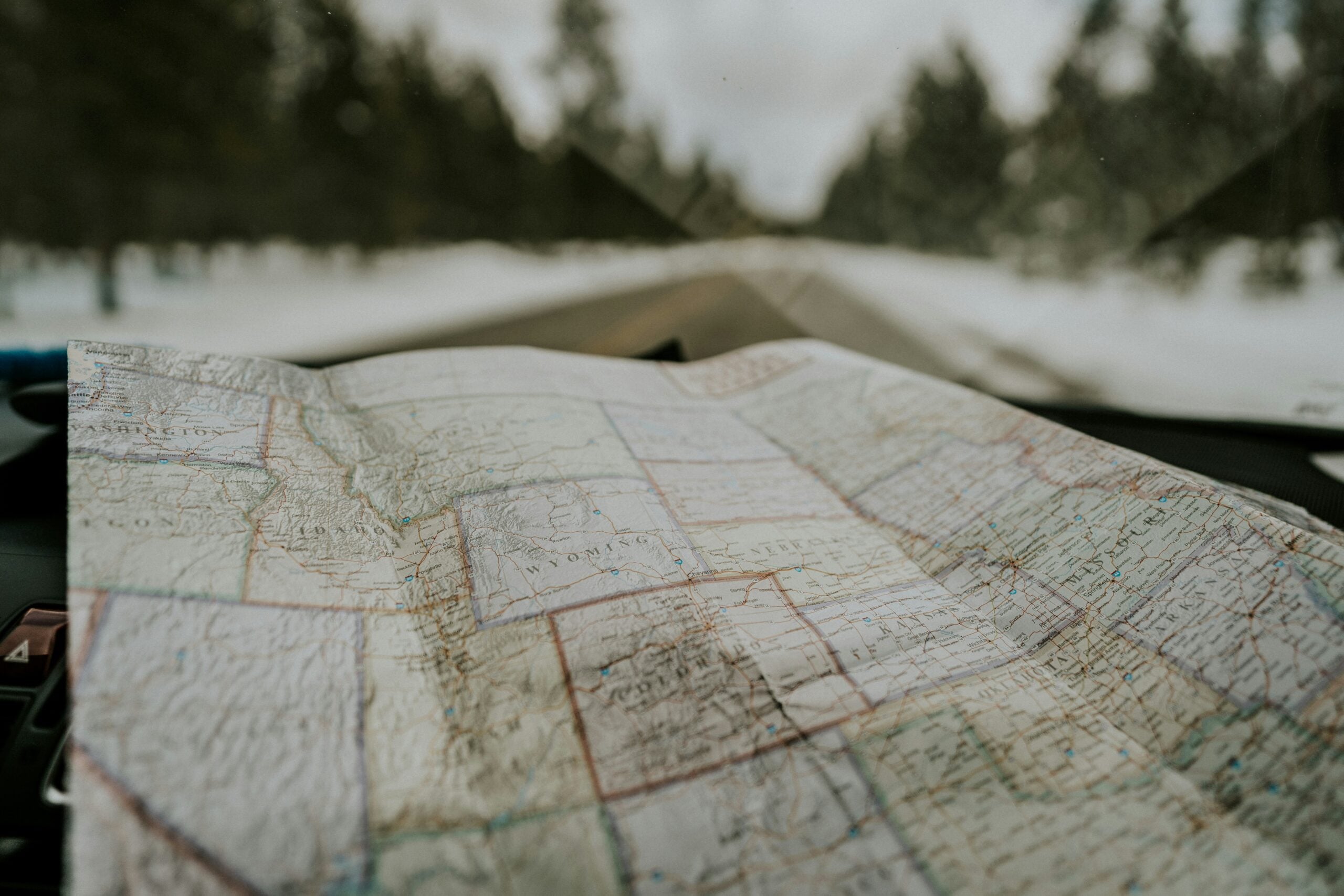 A paper map lies on a car dashboard, with a snowy road and pine trees visible through the windshield.