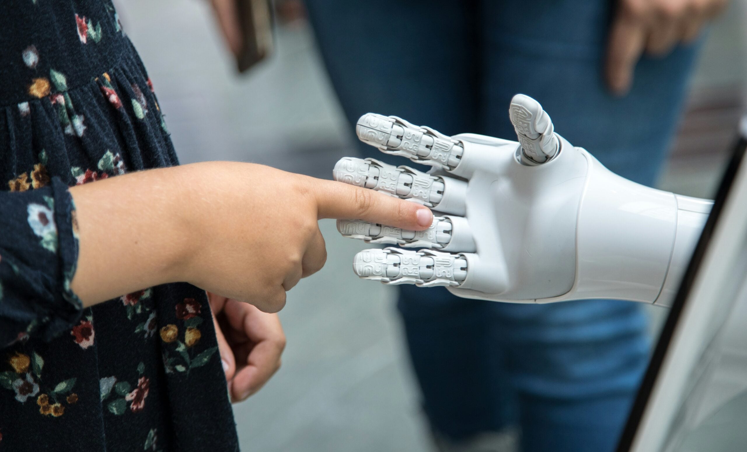 A child’s finger gently touches the fingertip of a white robotic hand, symbolizing human and technology connection—a poignant reflection of life in 2026 problems. The background shows blurred clothing and jeans, underscoring the everyday context in which these challenges play out.