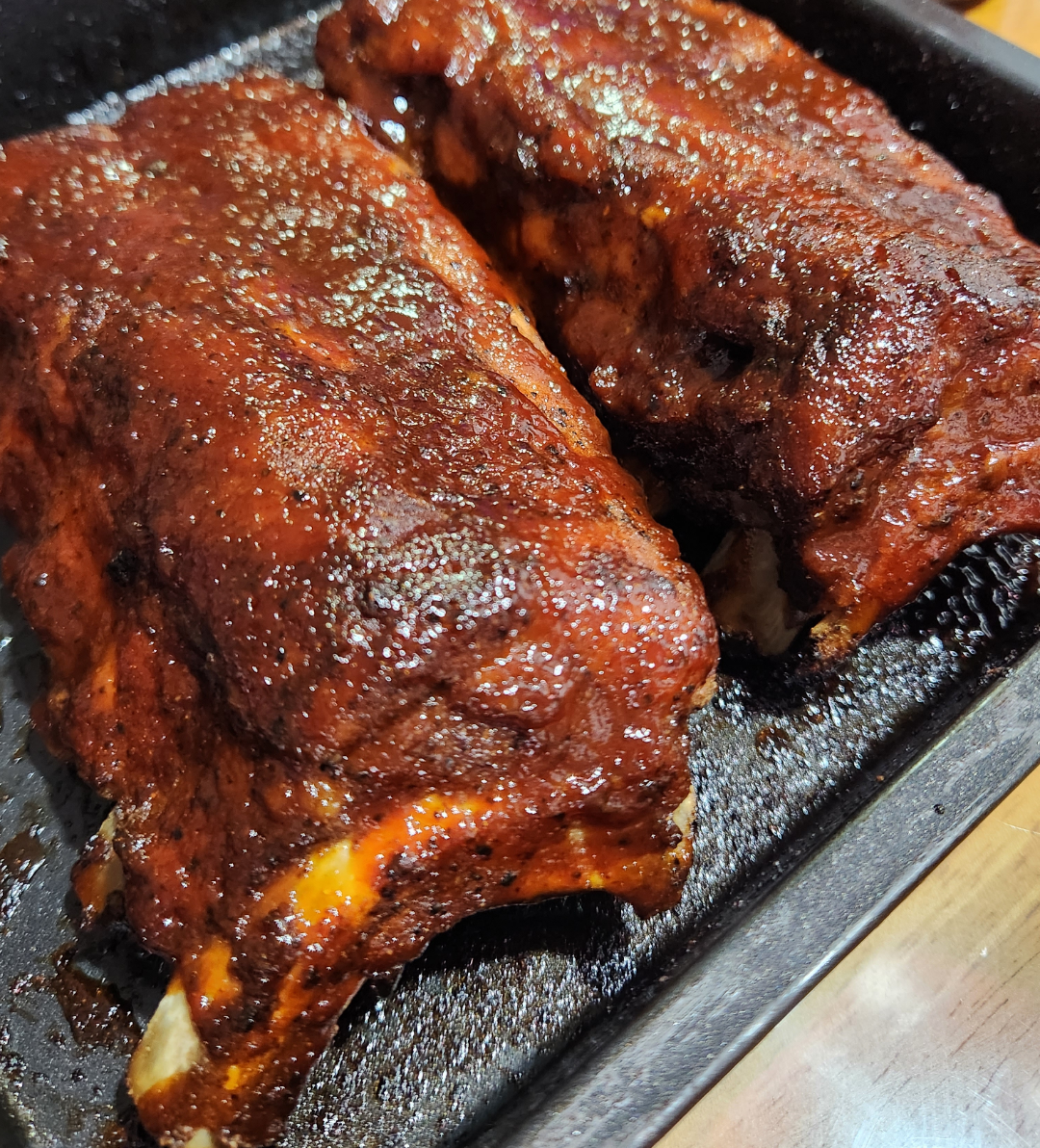 Two racks of barbecued ribs coated in a glossy, reddish-brown sauce rest in a black roasting pan on a wooden surface.