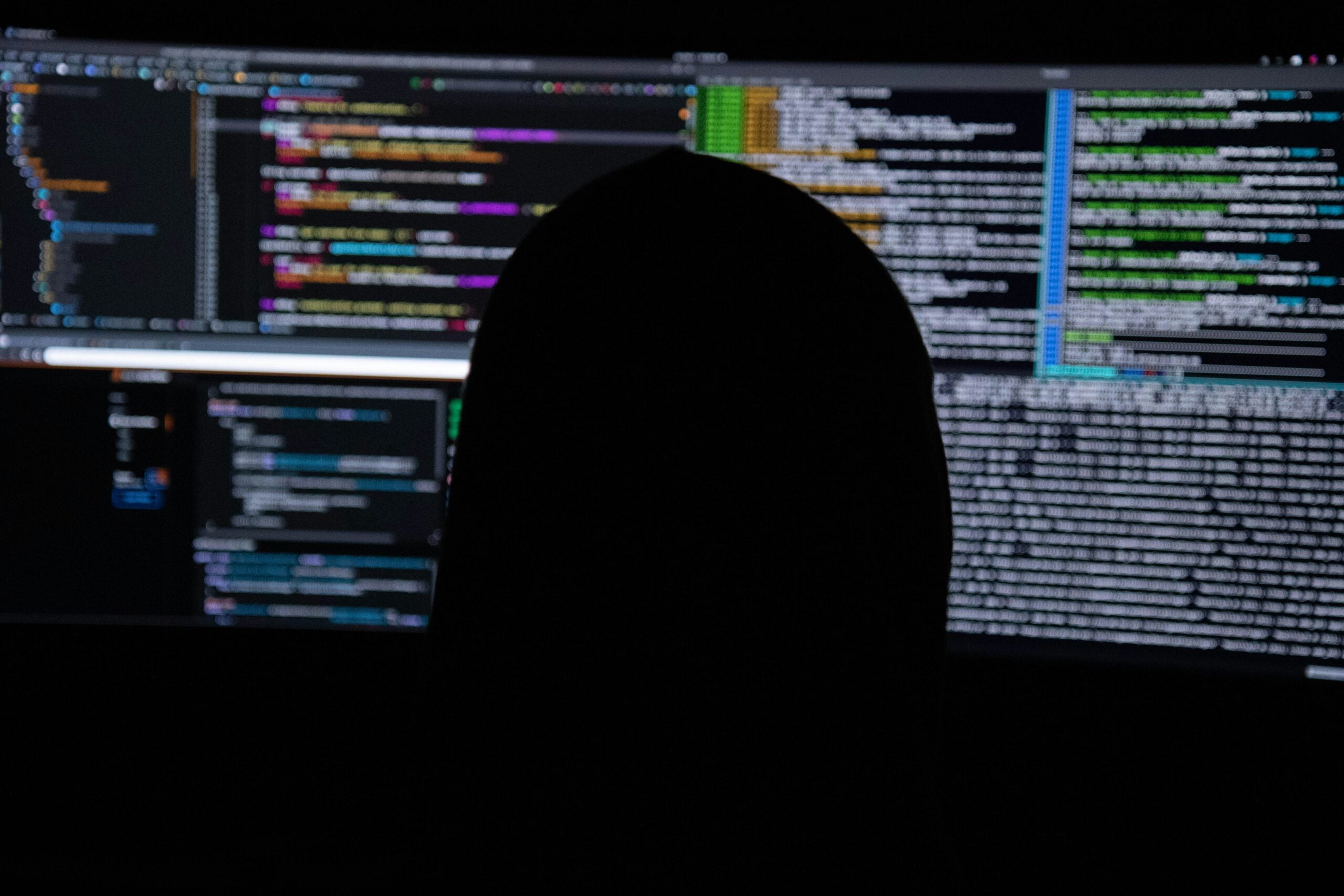 A person in silhouette sits in front of large computer monitors displaying colorful code and text on a dark background, suggesting programming or cybersecurity activity—perhaps tackling some of the complex life in 2026 problems that define our digital age.