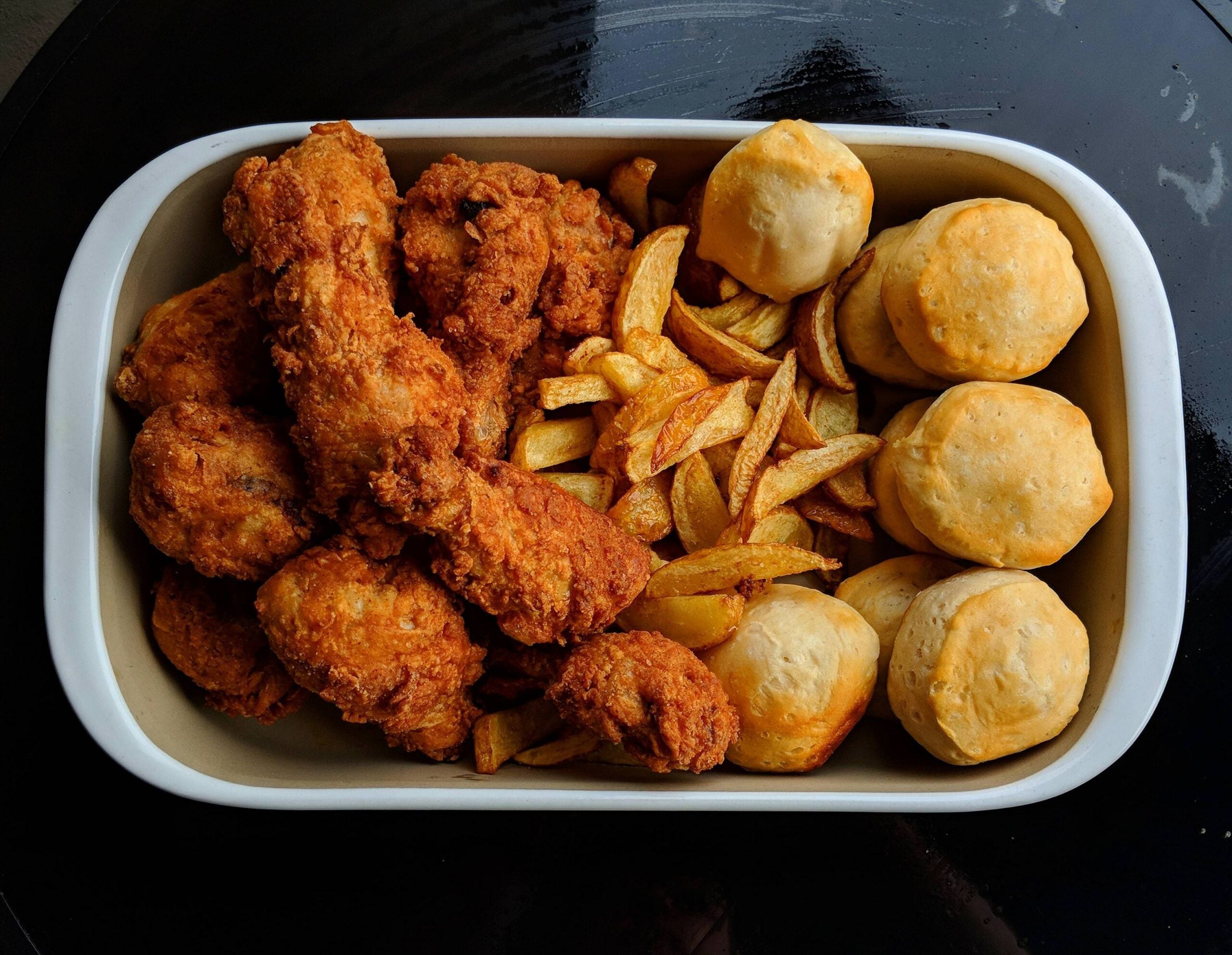 A white dish filled with fried chicken drumsticks, golden French fries, and round baked biscuits, all placed on a dark surface.