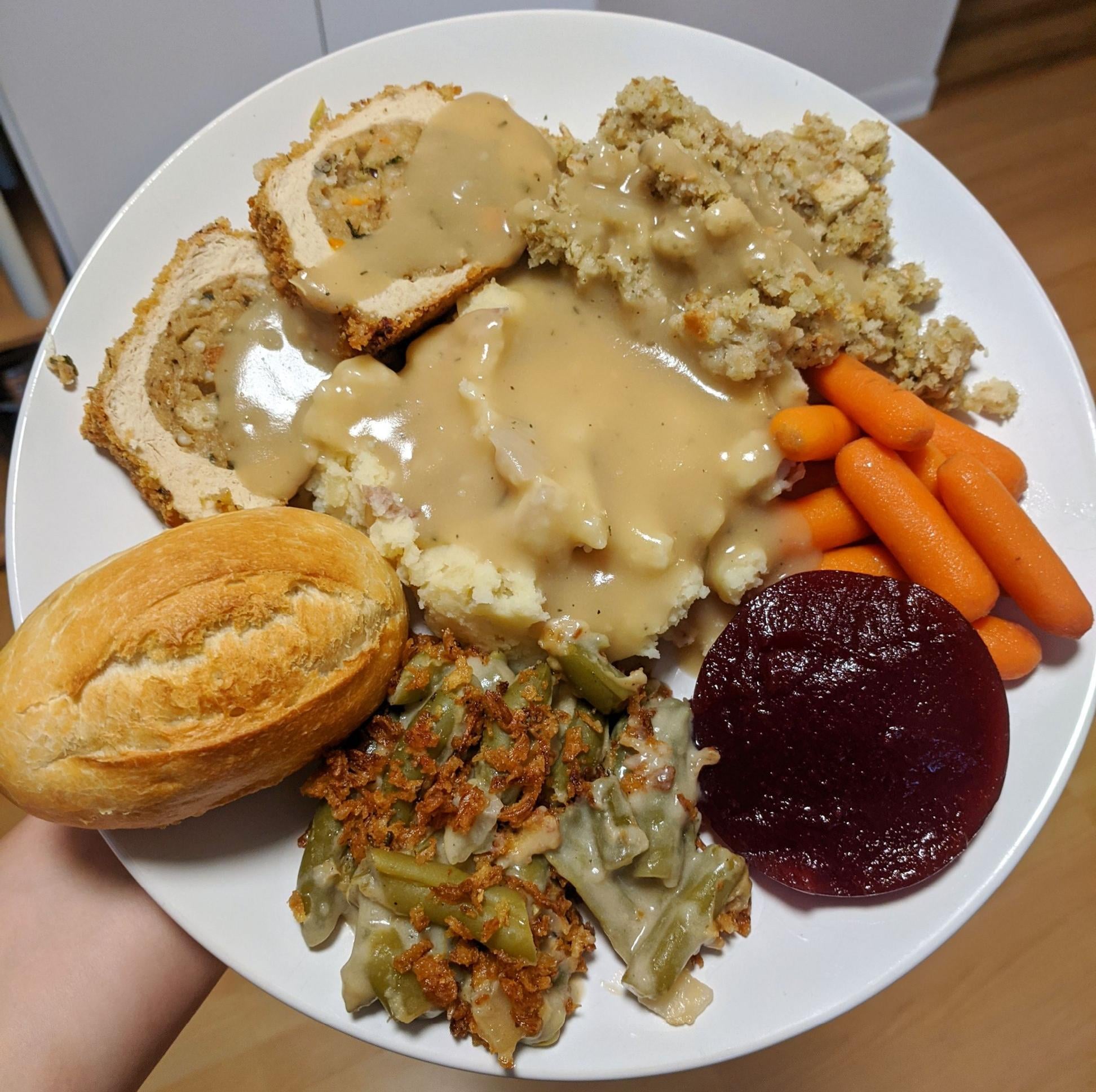 A plate of food with turkey and stuffing, mashed potatoes and gravy, baby carrots, cranberry sauce, green bean casserole, and a dinner roll.