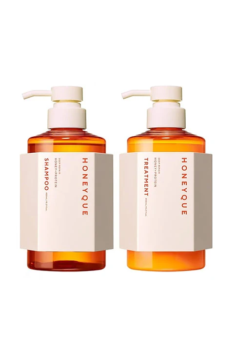 Two amber-colored pump bottles labeled “HONEYQUE.” One bottle is marked “Shampoo,” and the other “Treatment.” Both have white and beige labels with minimalistic design, standing side by side against a white background.