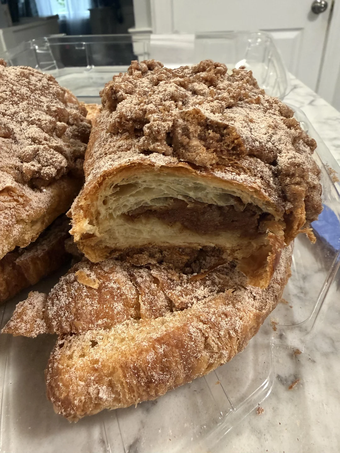 A close-up of sliced, flaky pastry with a crumbly, cinnamon-sugar topping and a visible cinnamon filling, placed on a marble countertop in a clear plastic container.