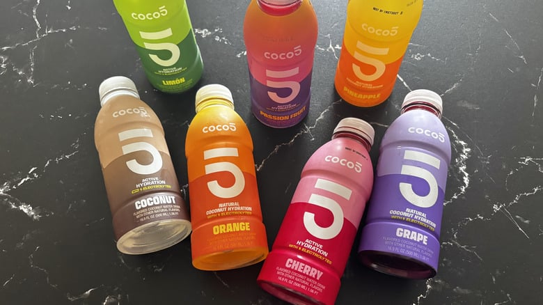 Seven bottles of Cocos 5 flavored coconut water in various colors and flavors, including lemon, pineapple, passion fruit, coconut, orange, cherry, and grape, arranged on a black marble surface.