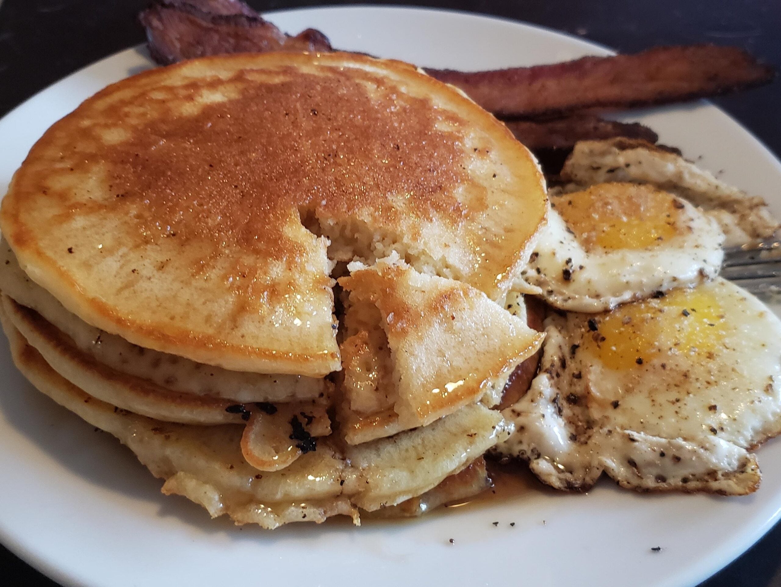 A stack of pancakes with syrup, two sunny-side-up eggs, and crispy bacon strips are served on a white plate.