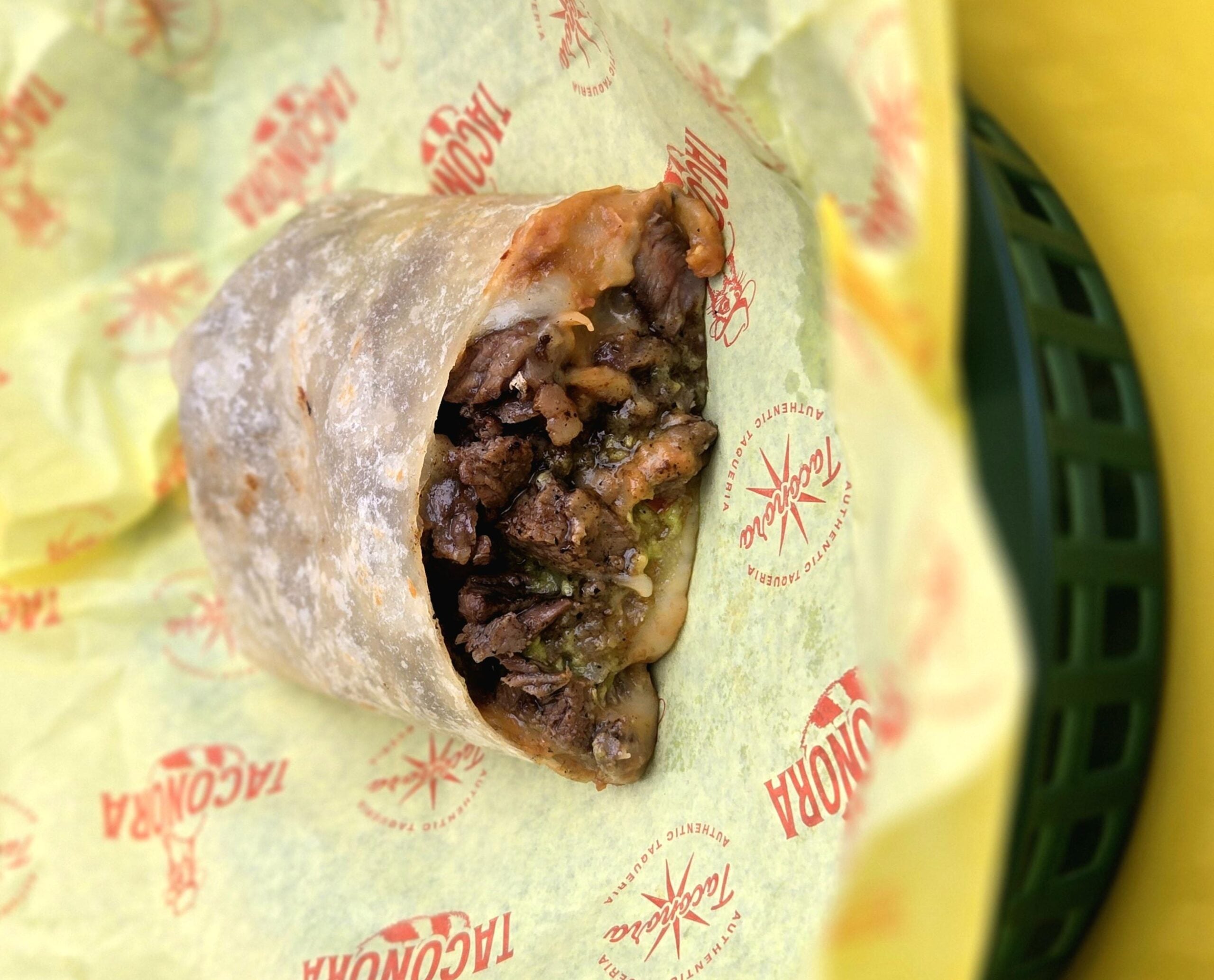 A close-up of a partially unwrapped burrito filled with grilled meat, melted cheese, and green salsa, sitting on yellow paper with “TACONAZO” branding inside a green basket.