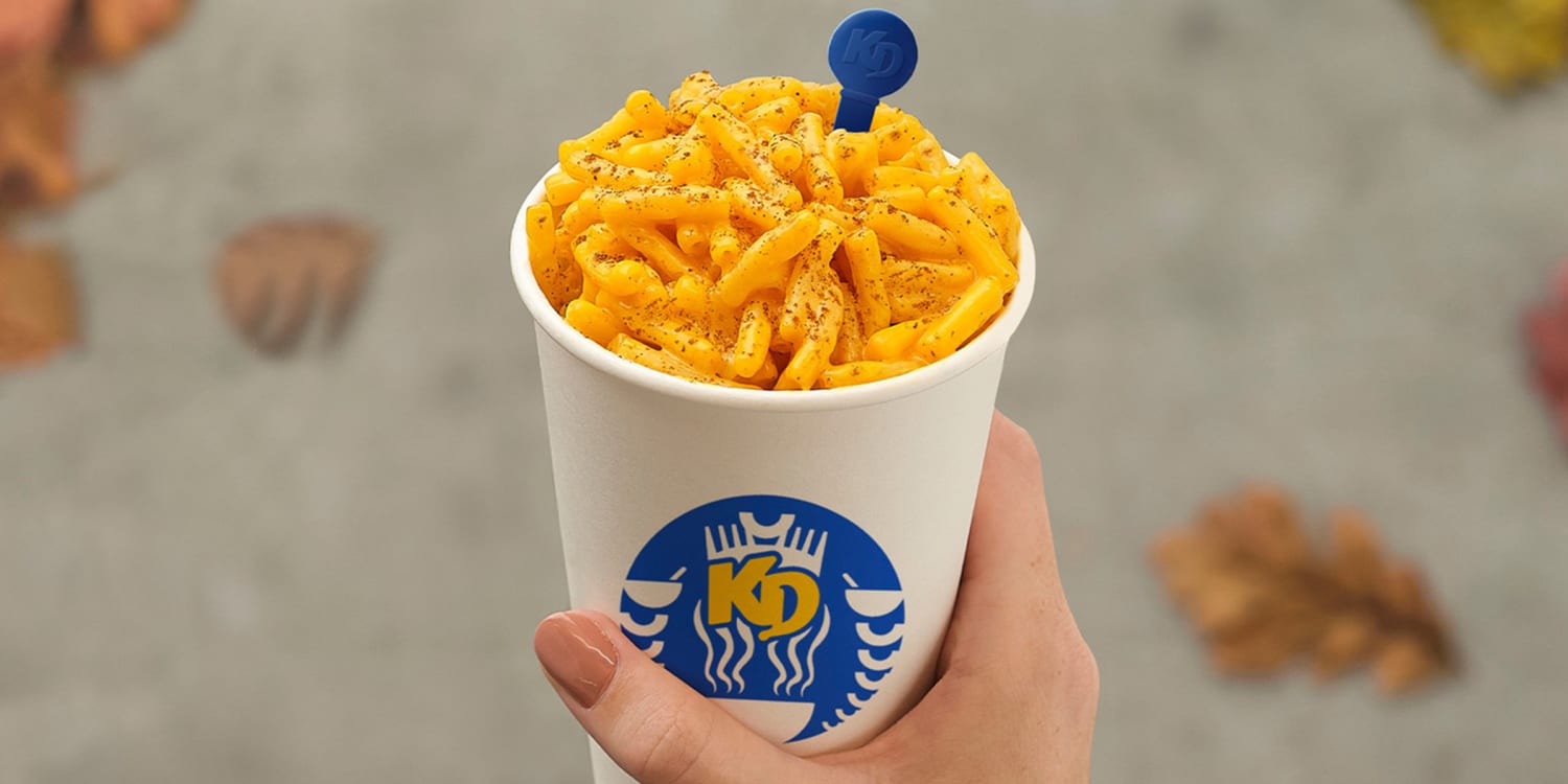 A hand holds a white cup with the KD logo, filled with macaroni and cheese topped with black pepper. Leaves are scattered on a blurred gray surface in the background.