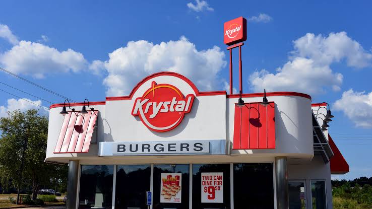 A Krystal fast-food restaurant with a red and white exterior, large Krystal logo, “BURGERS” sign, and promotional posters on the windows under a blue sky with clouds.