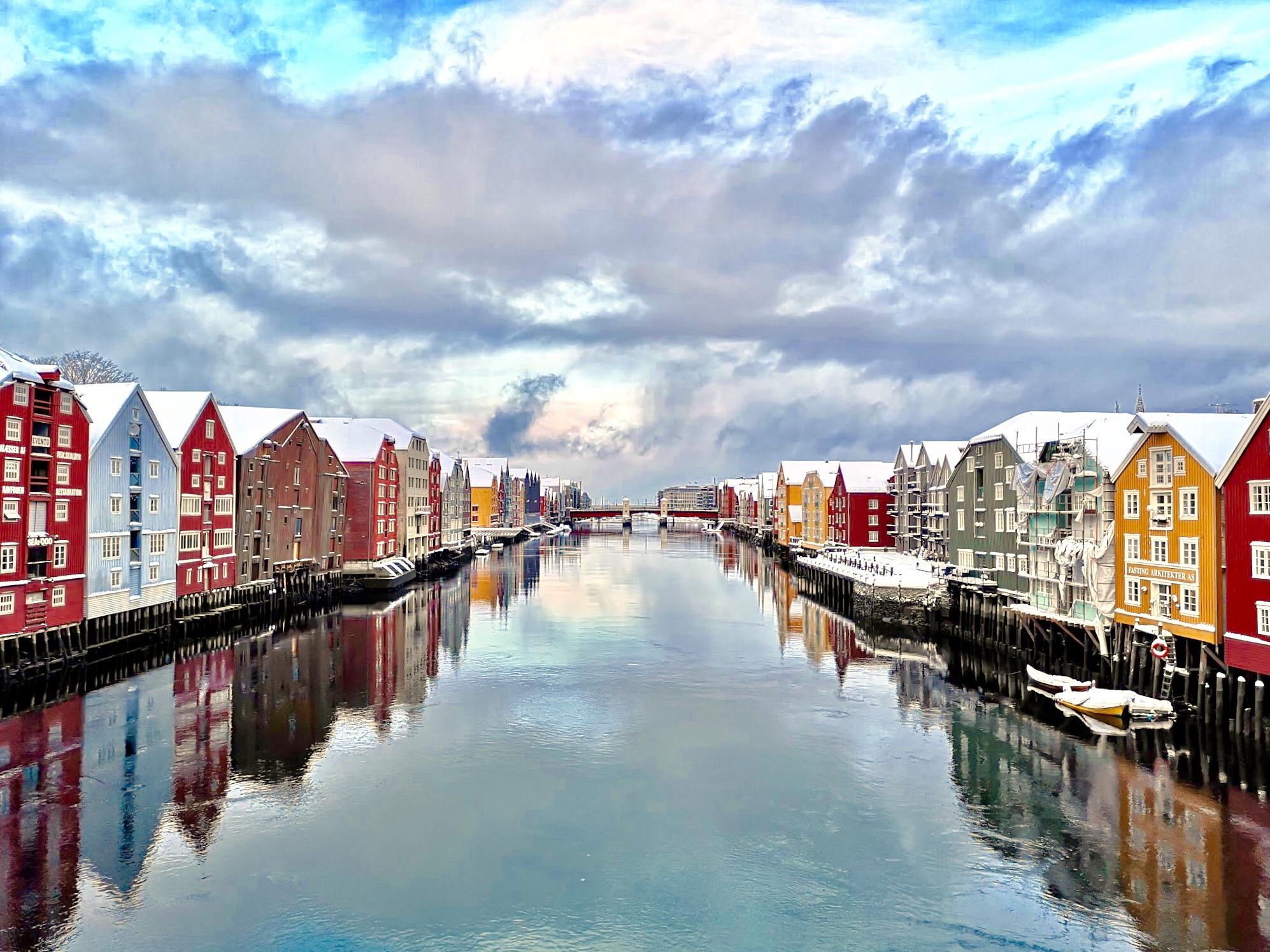 Colorful wooden buildings line both sides of a calm river under a cloudy sky, with their reflections visible in the water. Snow covers the rooftops, suggesting a winter day in a picturesque Nordic town.