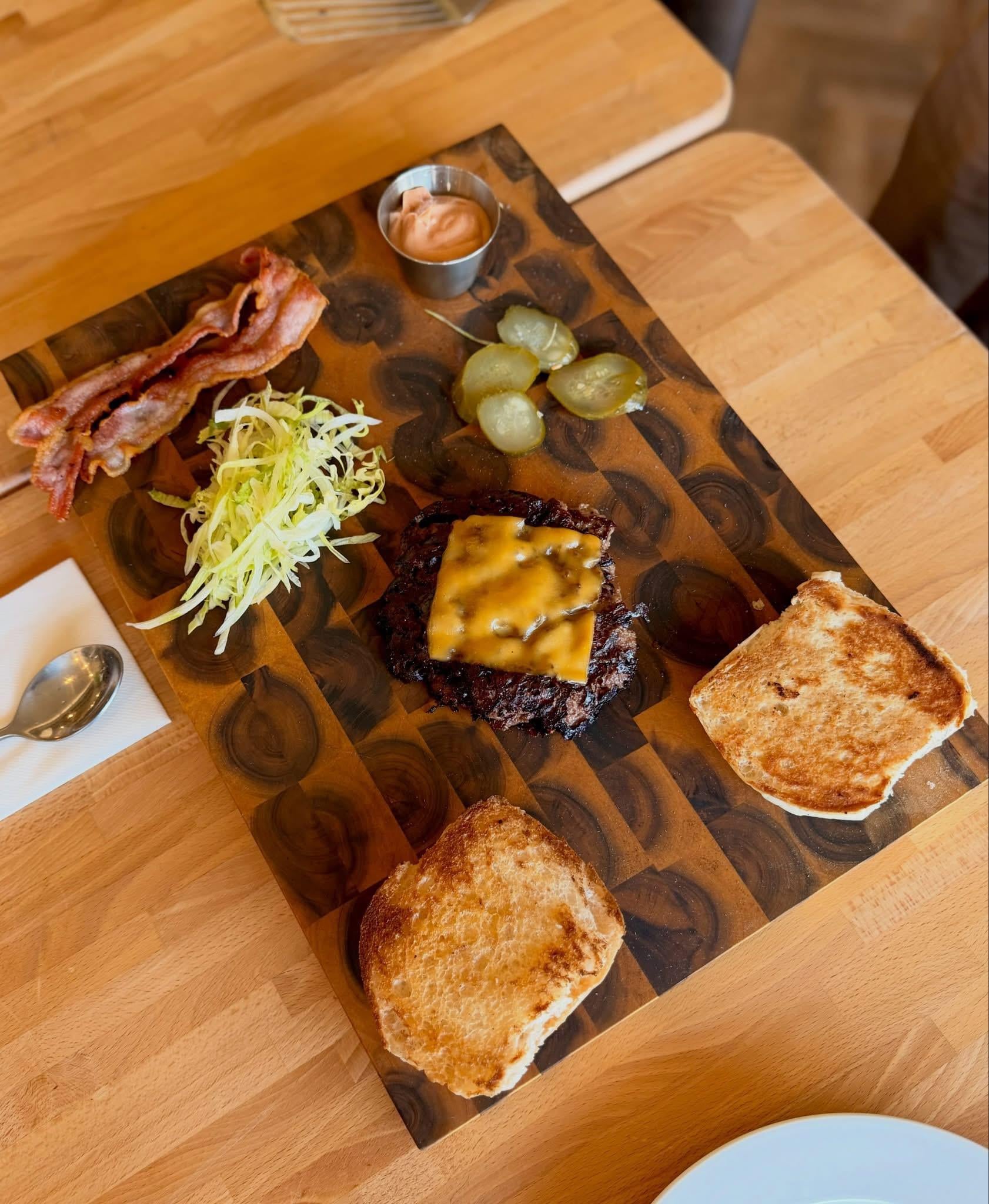 A wooden board with a grilled burger patty topped with melted cheese, toasted burger buns, crispy bacon strips, shredded lettuce, green pickles, and a small cup of dipping sauce on a wooden table.