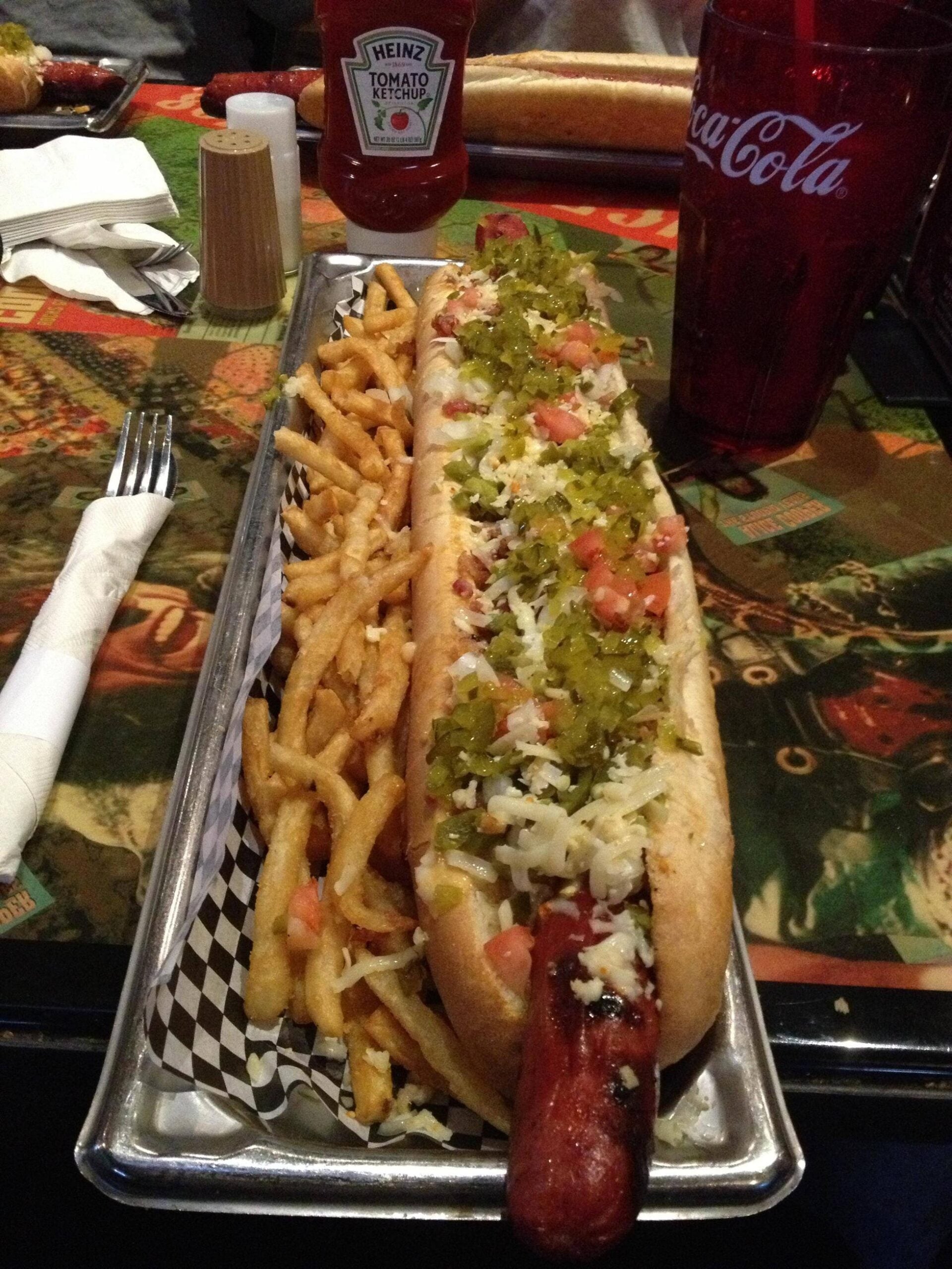 A long hot dog topped with relish, diced tomatoes, and shredded cheese sits next to a pile of fries on a tray. A fork, napkin, ketchup bottle, and Coca-Cola cup are on the colorful table.