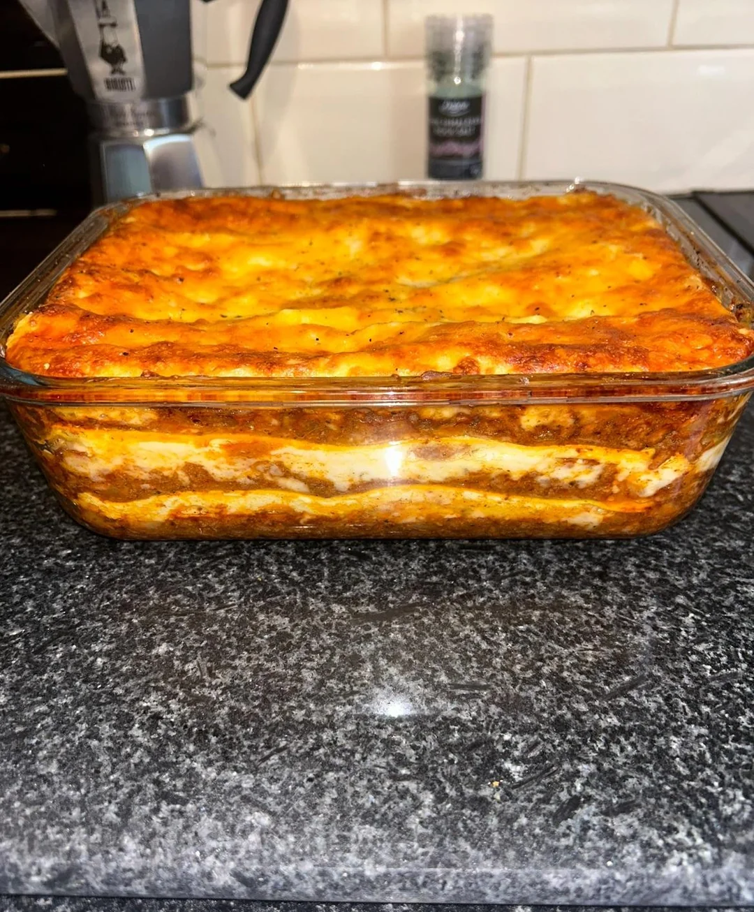 Homemade lasagna. A glass dish filled with baked lasagna shows visible layers of pasta, cheese, and meat sauce, sitting on a dark granite countertop.