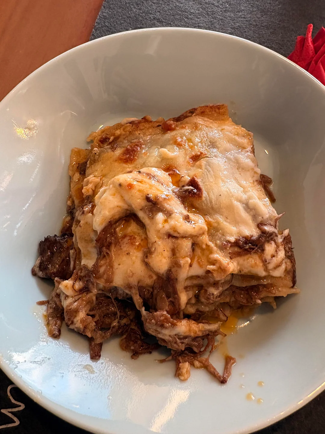 A slice of lasagna with melted cheese and meat sauce is served on a white plate. The lasagna looks creamy and rich, with visible layers and a browned top.