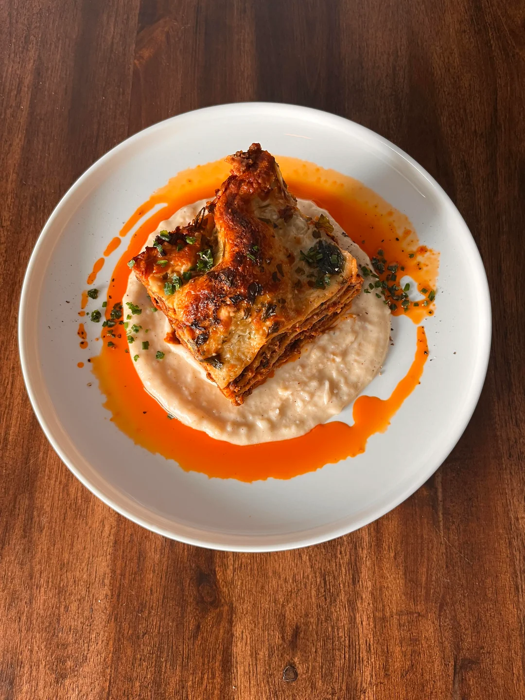 A slice of lasagna sits atop a creamy white sauce, garnished with chopped herbs and surrounded by a ring of orange oil on a white plate with a wooden table background.