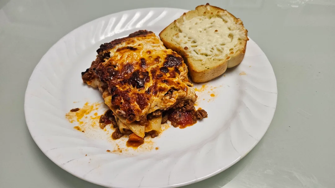A serving of lasagna with layers of cheese, sauce, and meat is on a white plate next to a slice of toasted garlic bread with melted cheese on top.